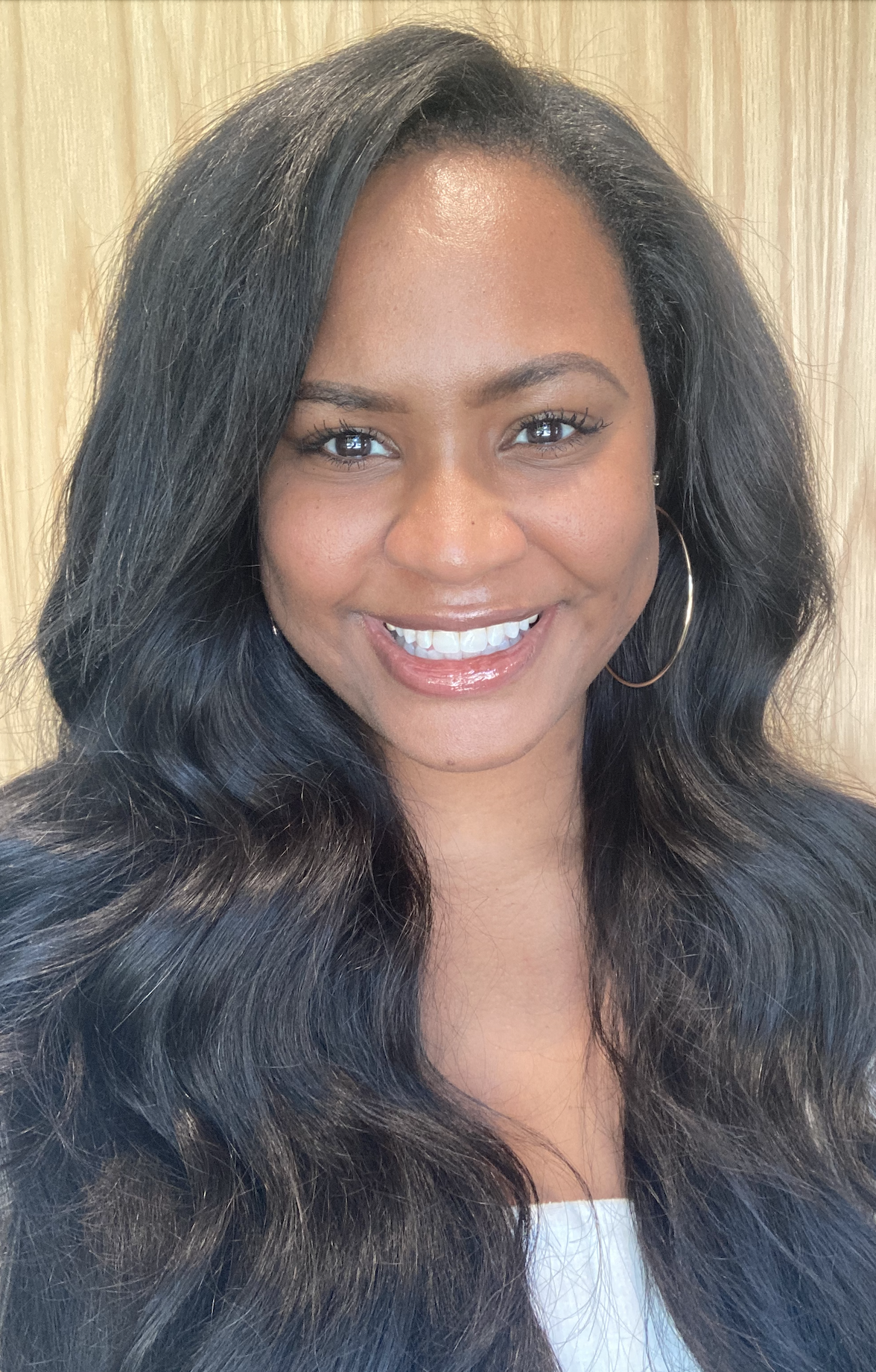 A smiling woman with long, wavy black hair, wearing hoop earrings, standing in front of a wooden background.