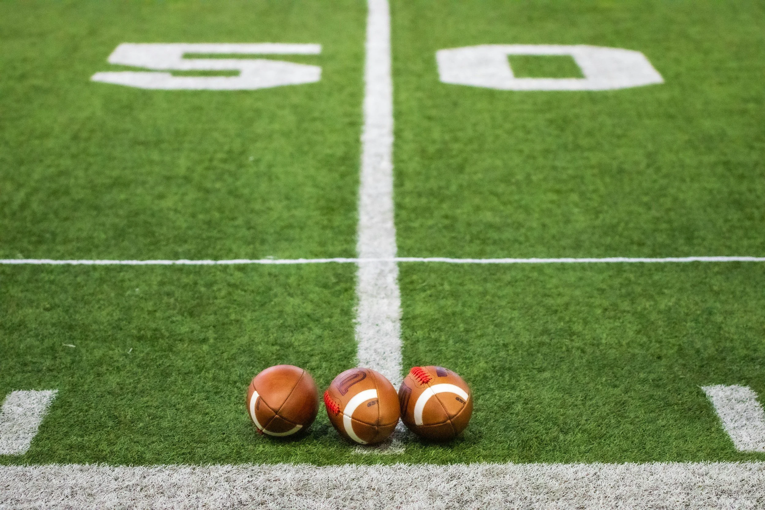 Three footballs placed on the sideline of a football field with green artificial turf, white yard line markings, and the number 50 visible in the background.