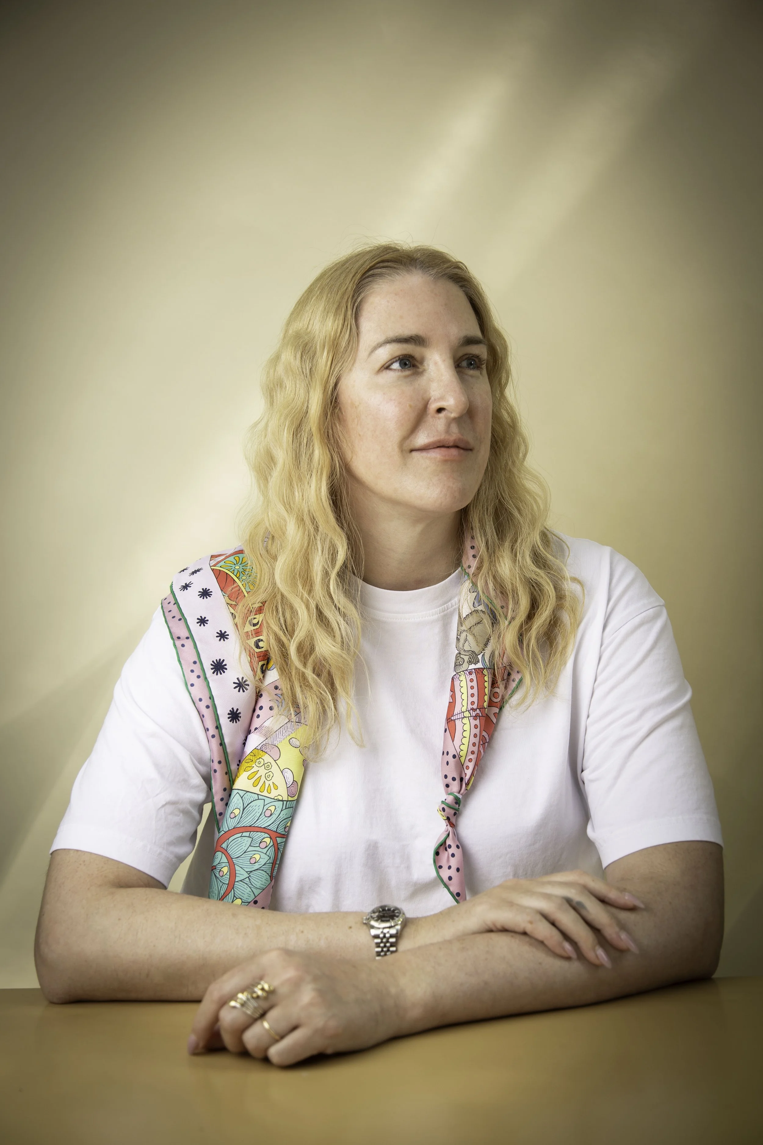 A woman with blonde, wavy hair sitting at a table, wearing a white t-shirt and a colorful scarf around her neck, with a neutral-colored background.