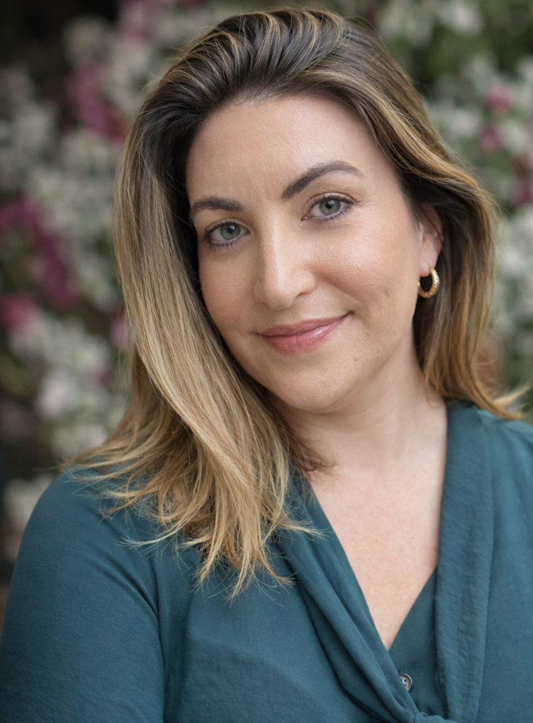 Close-up of a woman with shoulder-length blonde hair, wearing a teal blouse and gold hoop earrings, smiling softly outdoors with blurred flowers in the background.