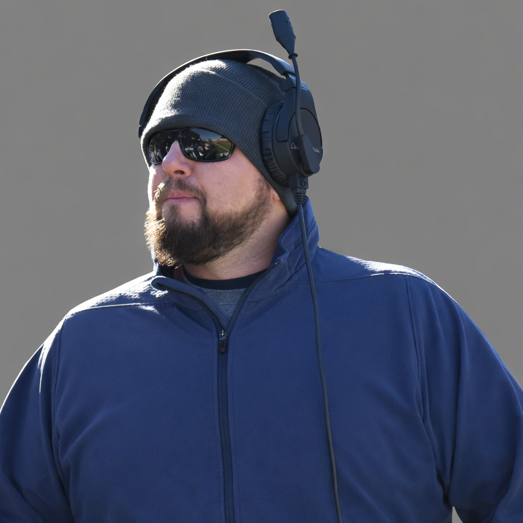 A man wearing black sunglasses, a gray beanie, and a blue jacket with a zipper, and a coaching headset.