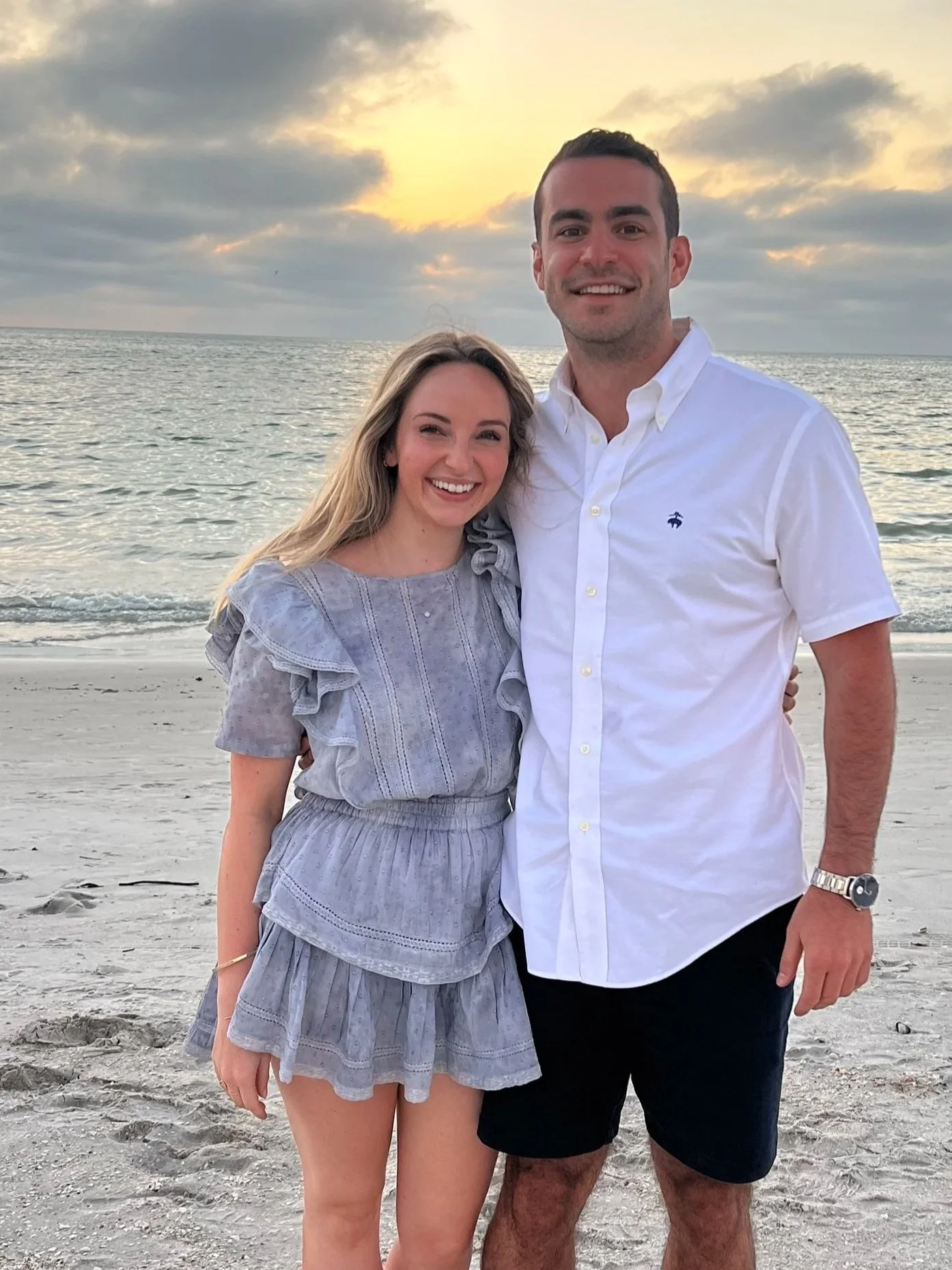 Ben and Anna, Crown Estate Management co-founders, stand on a sandy beach at sunset, with the ocean and cloudy sky in the background.