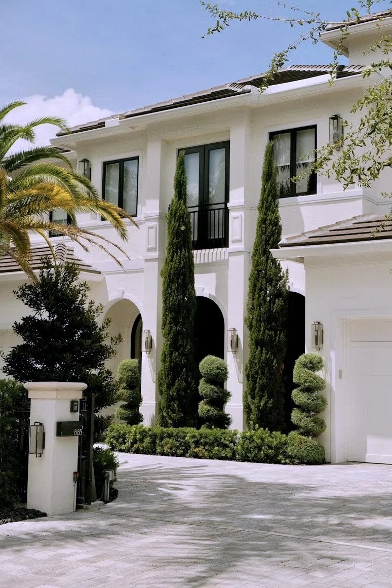 Front view of a modern white house with tall, narrow cypress trees and neatly trimmed bushes lining the pathway leading to the entrance.