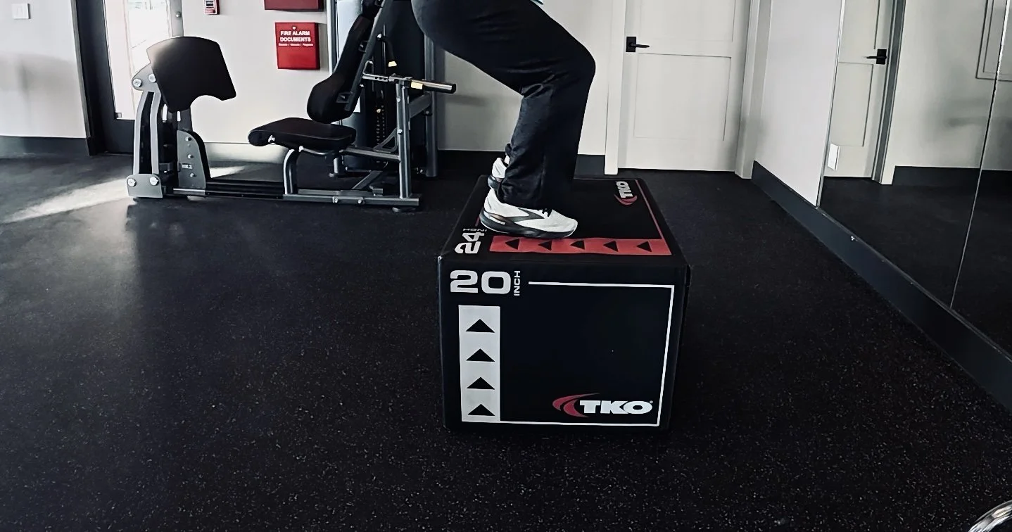 A person standing on a black and red TKO plyometric box in a gym, with a black exercise bench and a fire alarm on the wall in the background.