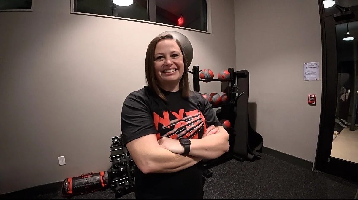 Woman smiling with arms crossed in a gym with fitness equipment behind her.