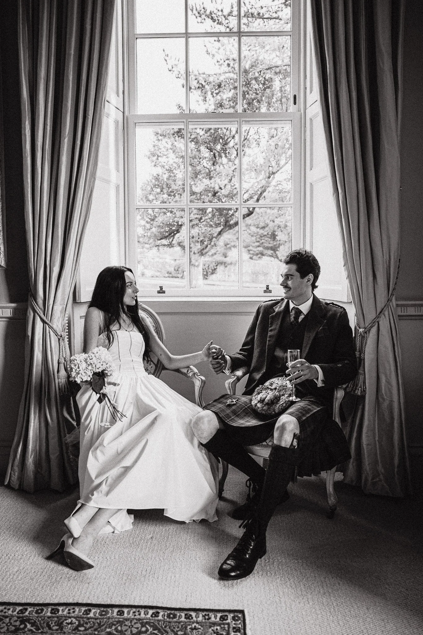 A black-and-white photo of a bride and groom sitting on a window seat, holding hands and smiling at each other. The bride is in a strapless wedding gown holding a bouquet, and the groom is in a kilt with a jacket, holding a glass of champagne. The large window behind them shows trees outside, framed by curtains.
