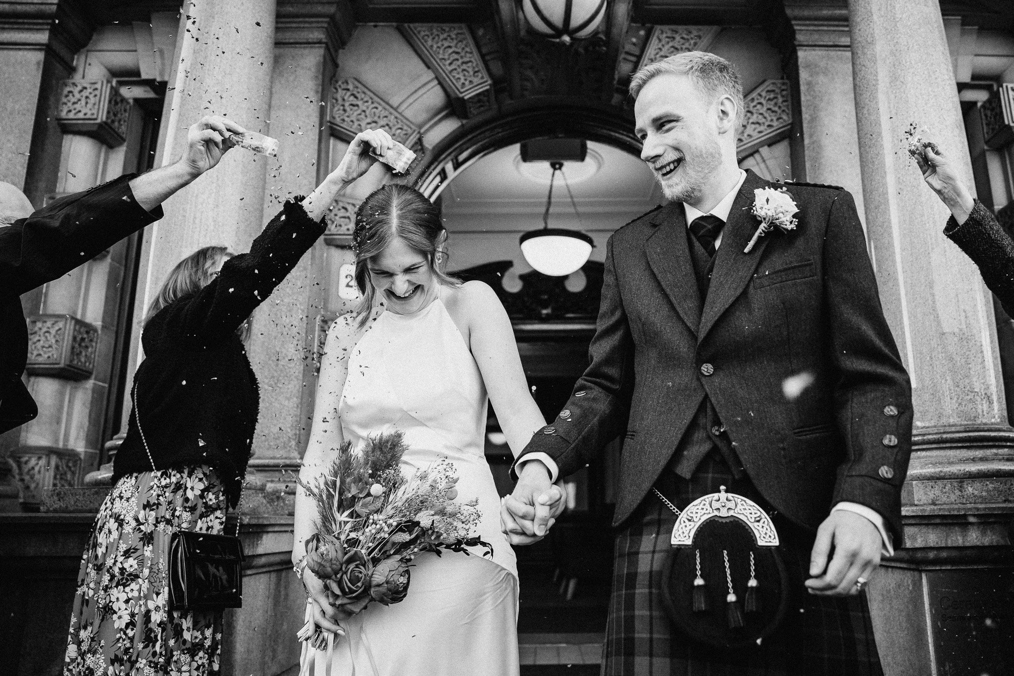 A bride and groom exiting a building with people celebrating around them, some throwing confetti, in black and white.
