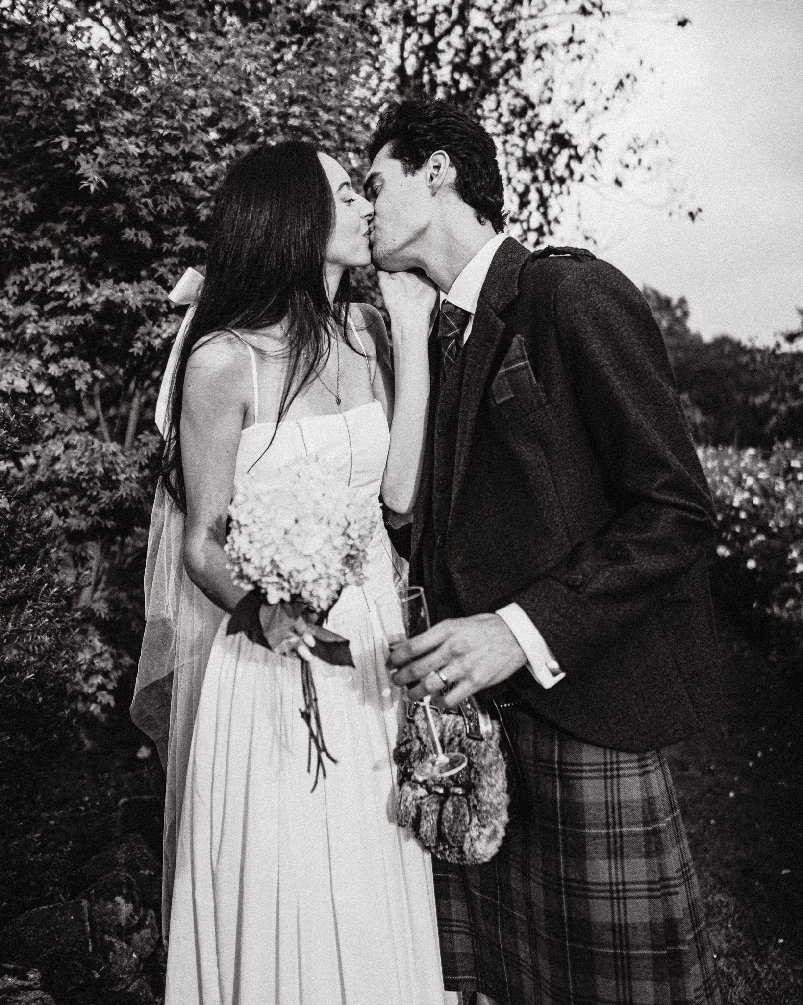 A black and white photo of a couple sharing a kiss outdoors at their wedding, with the woman holding a bouquet and the man holding a drink.