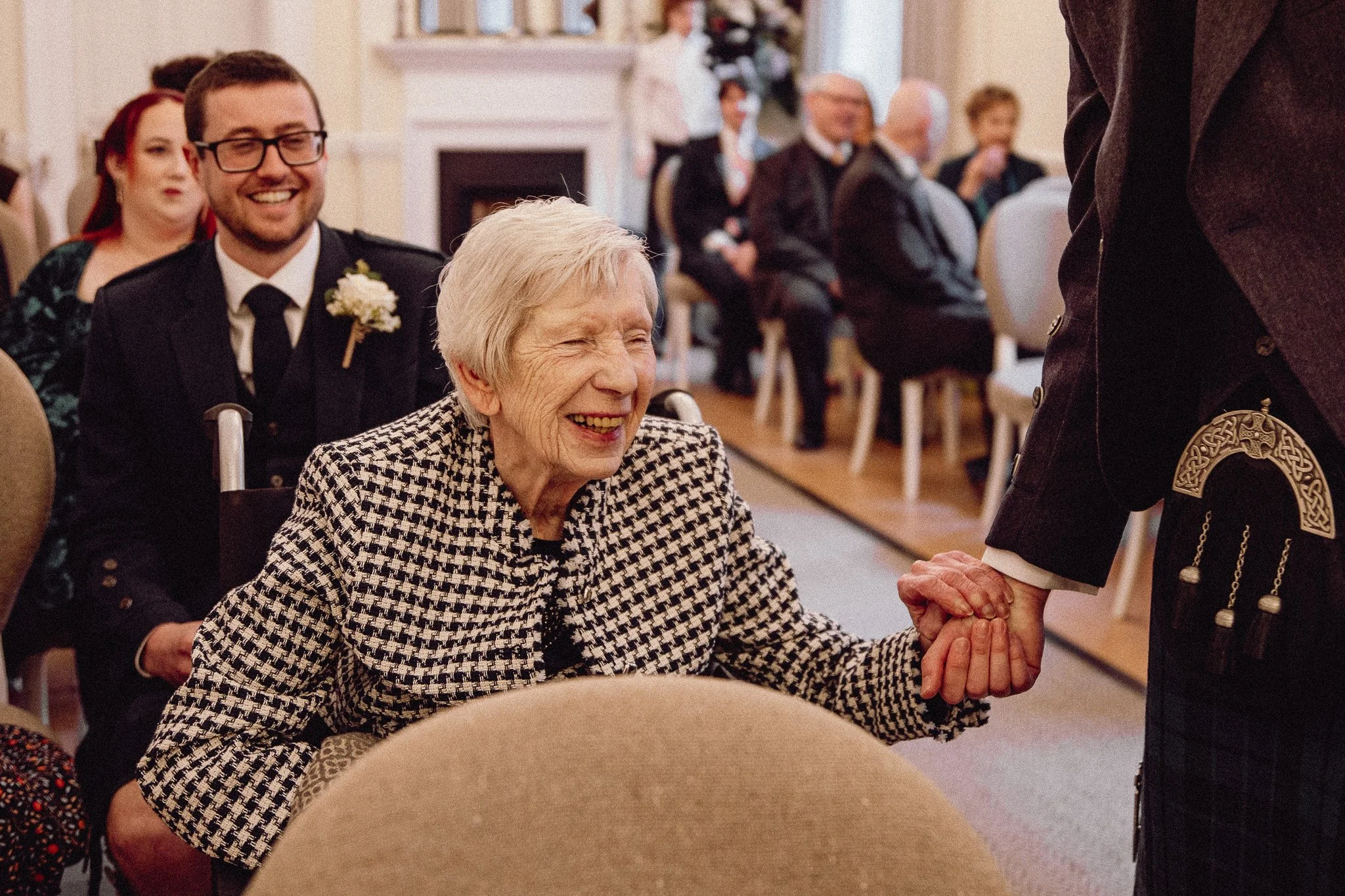 An elderly woman with short grey hair, smiling and holding hands with a man dressed in traditional Scottish attire, at a formal event or celebration in a decorated room.