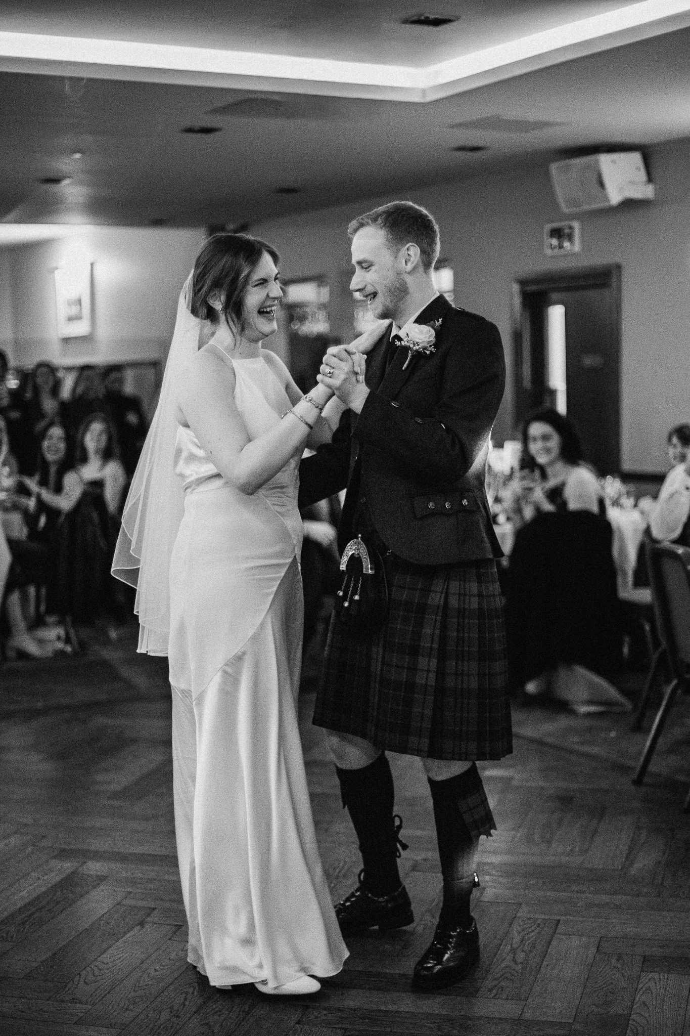 Black and white photo of a bride and groom dancing and smiling at their wedding reception, with guests seated at tables in the background.