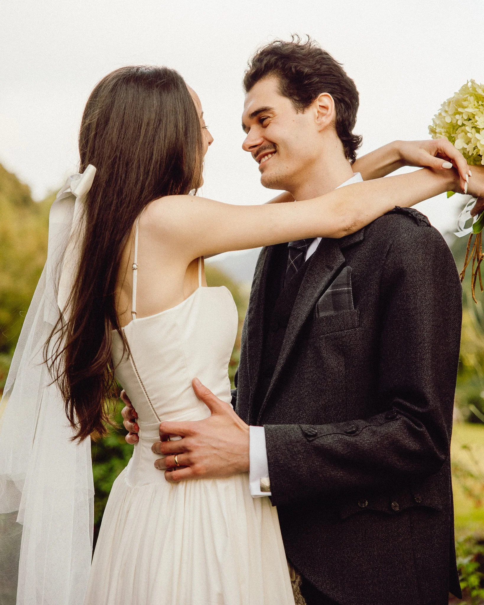 A bride and groom hugging outdoors during their wedding, smiling at each other, with the bride holding a bouquet of white flowers.