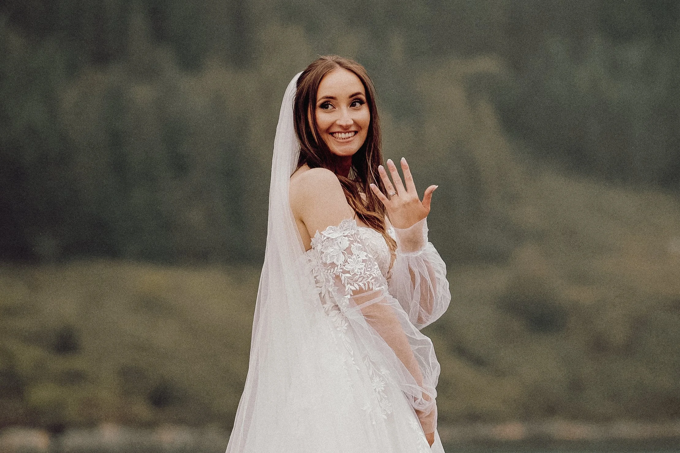 A smiling bride in a wedding dress outdoors, showing her wedding ring to camera with her right hand.