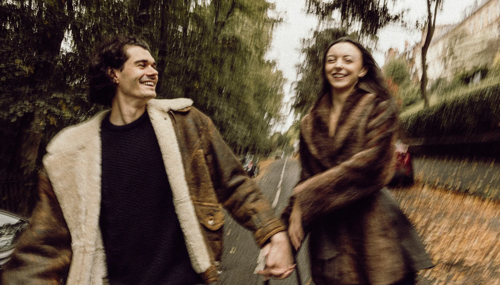 A man and woman holding hands and smiling, walking outdoors on a street with trees and fallen leaves.