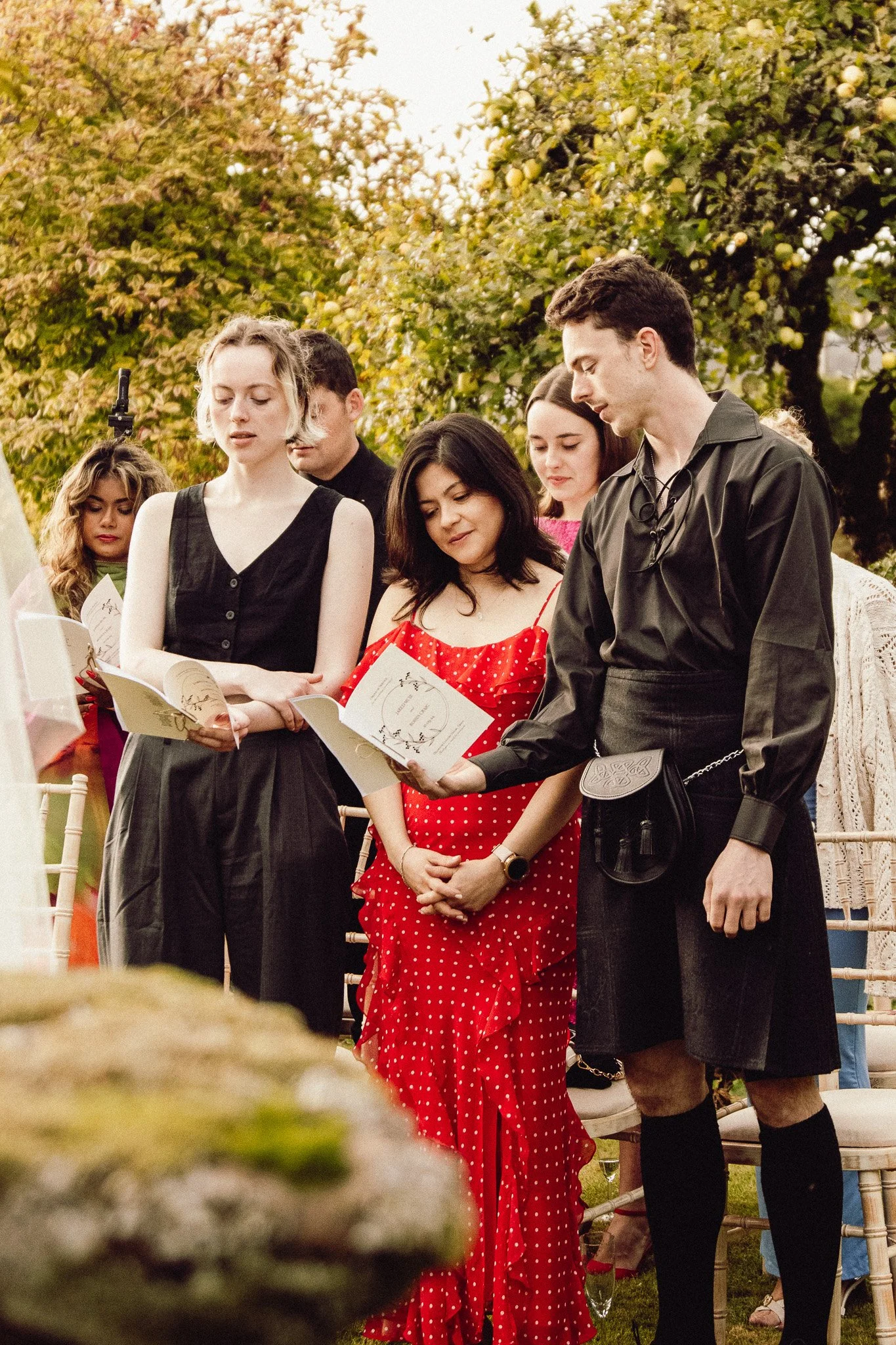 People gathered outdoors, reading from booklets during a ceremony or event, with trees in the background and autumn foliage.