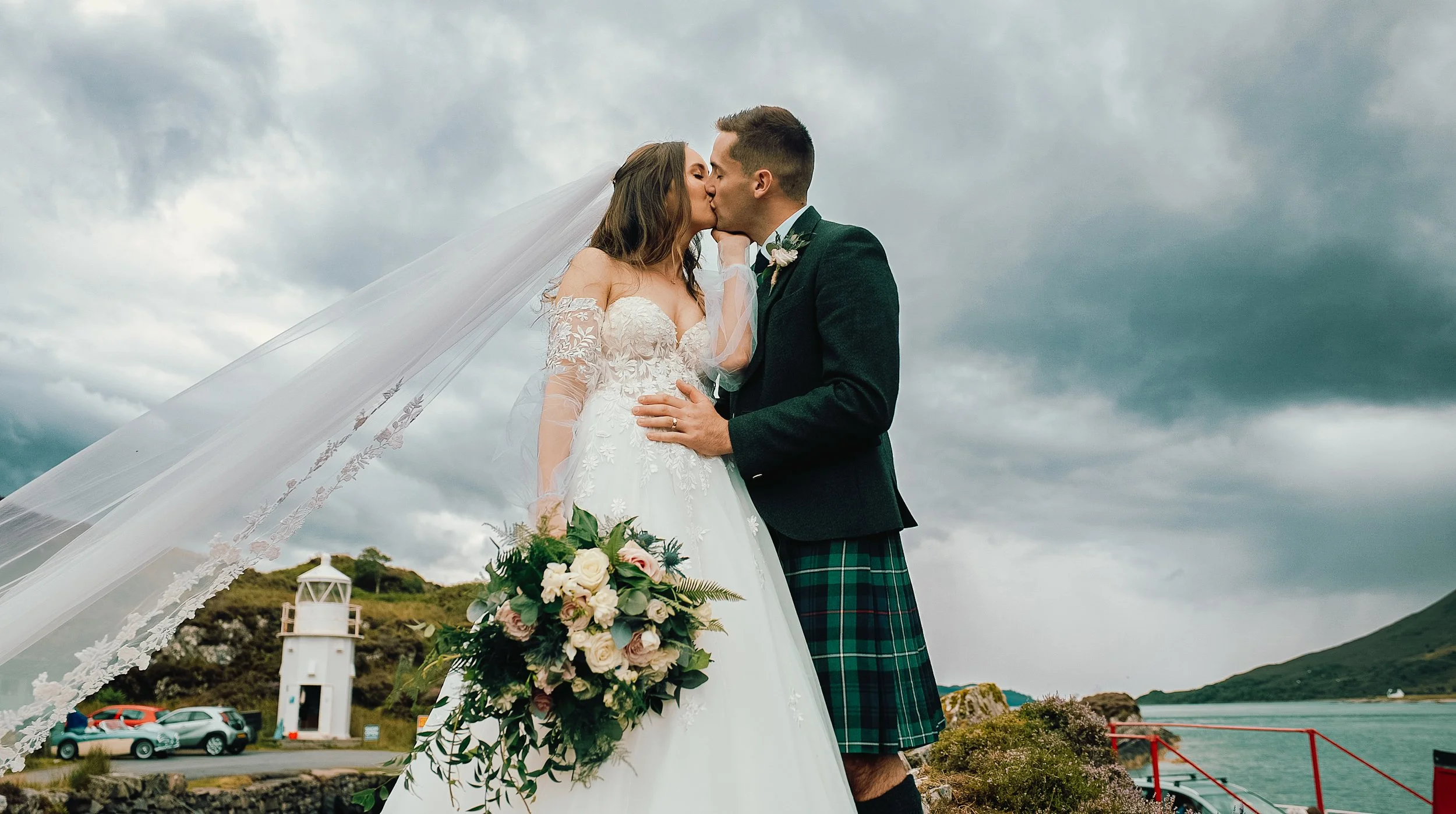 A bride and groom kiss outdoors near a body of water with a cloudy sky. The bride wears a white wedding gown and veil, holding a bouquet of white and pink flowers. The groom wears a traditional kilt with a dark jacket and a boutonniere.