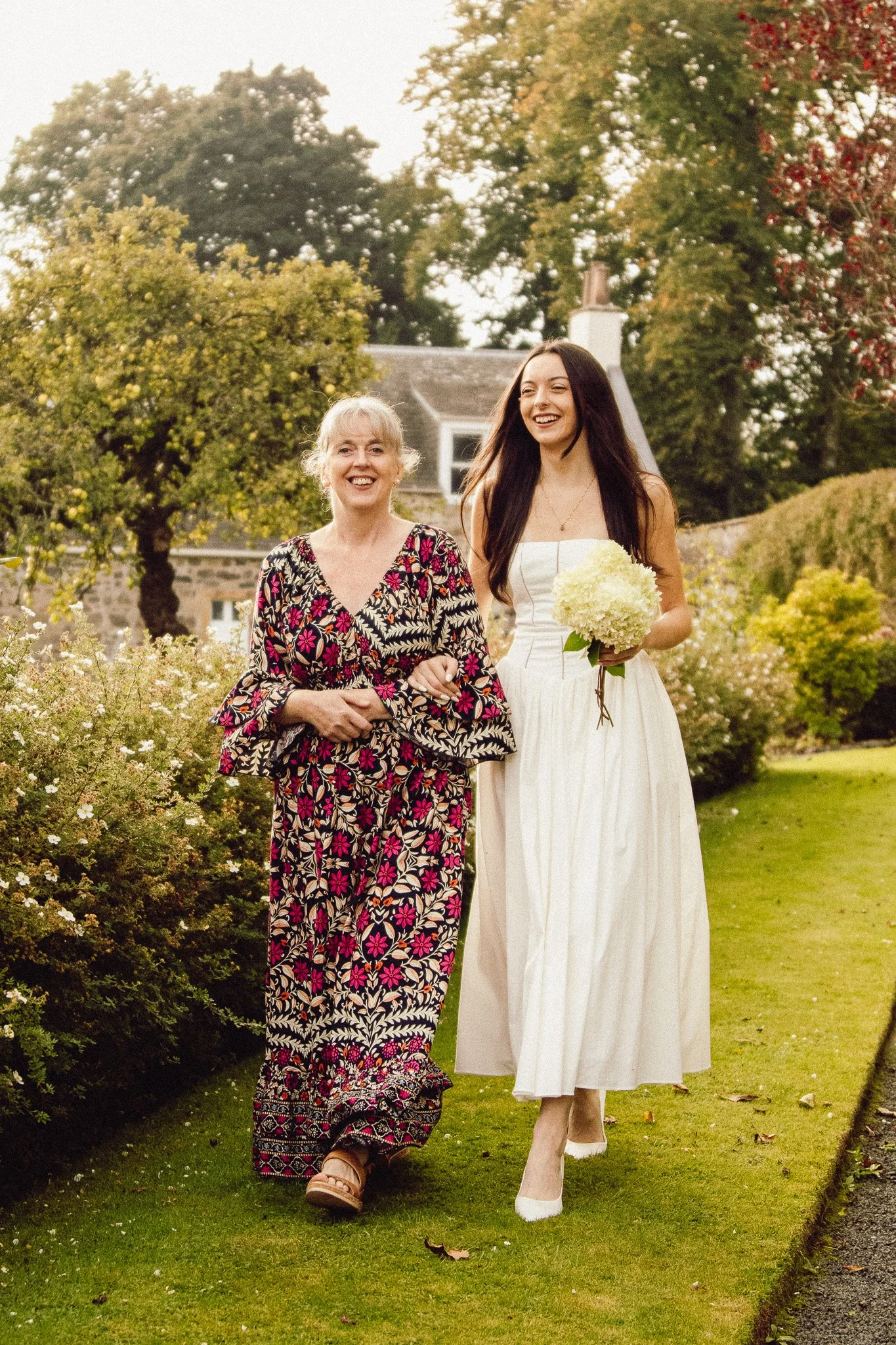 A young woman in a white dress holding a bouquet walking with an older woman in a colorful floral dress outdoors on a grassy path with trees and a house in the background.