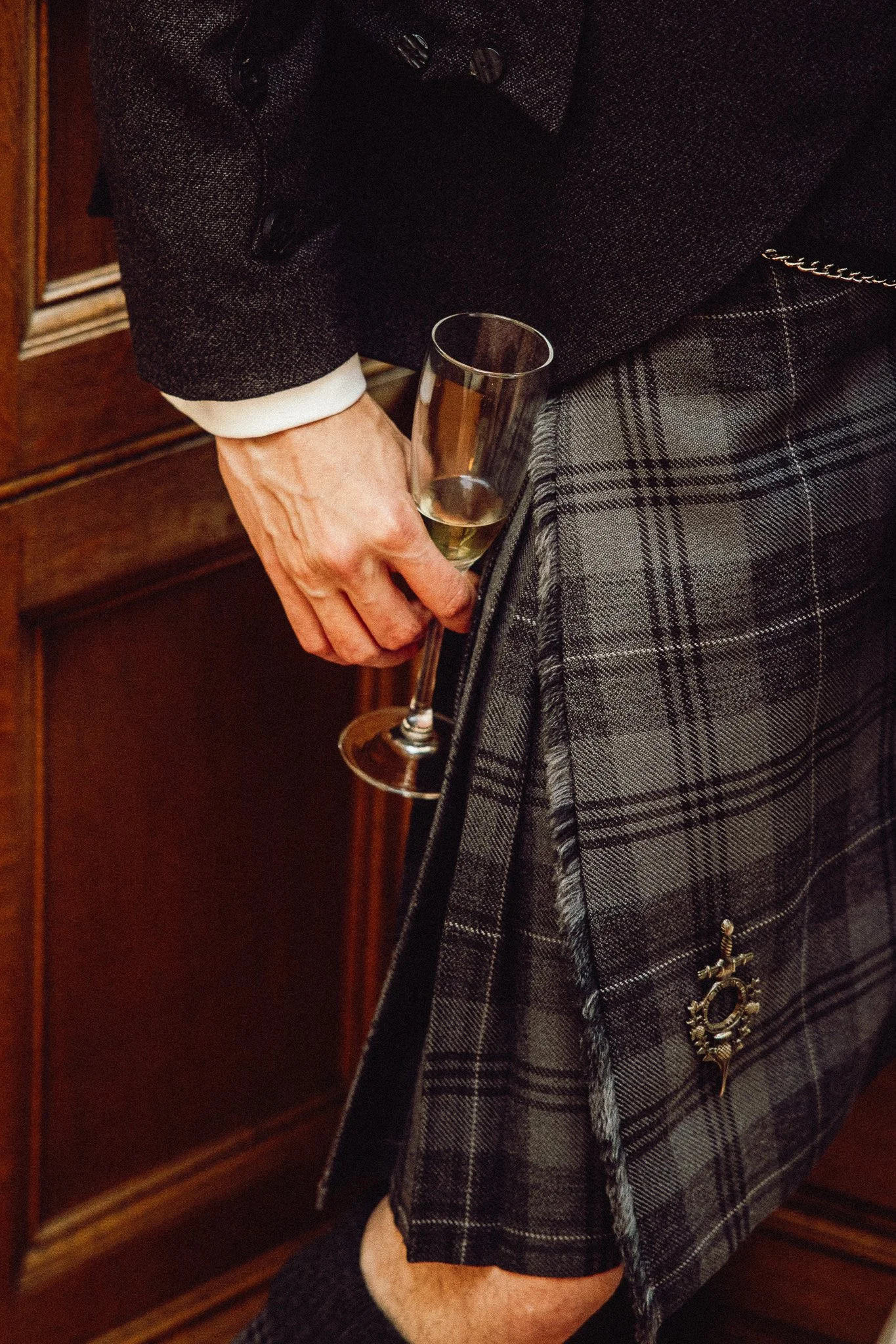 A person dressed in traditional Scottish attire, holding a glass of champagne, with a tartan kilt and a brooch