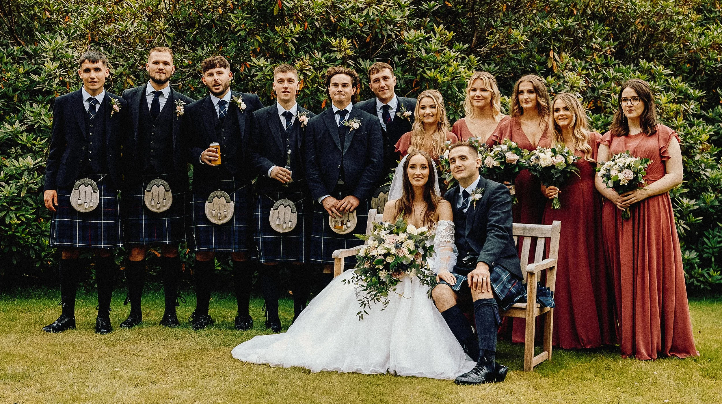 A wedding party outdoors on grass in front of shrubbery. The bride and groom are seated in the center, with the bride in a white wedding dress holding a large bouquet, and the groom in a dark suit with a kilt. Behind them are eight men dressed in dar