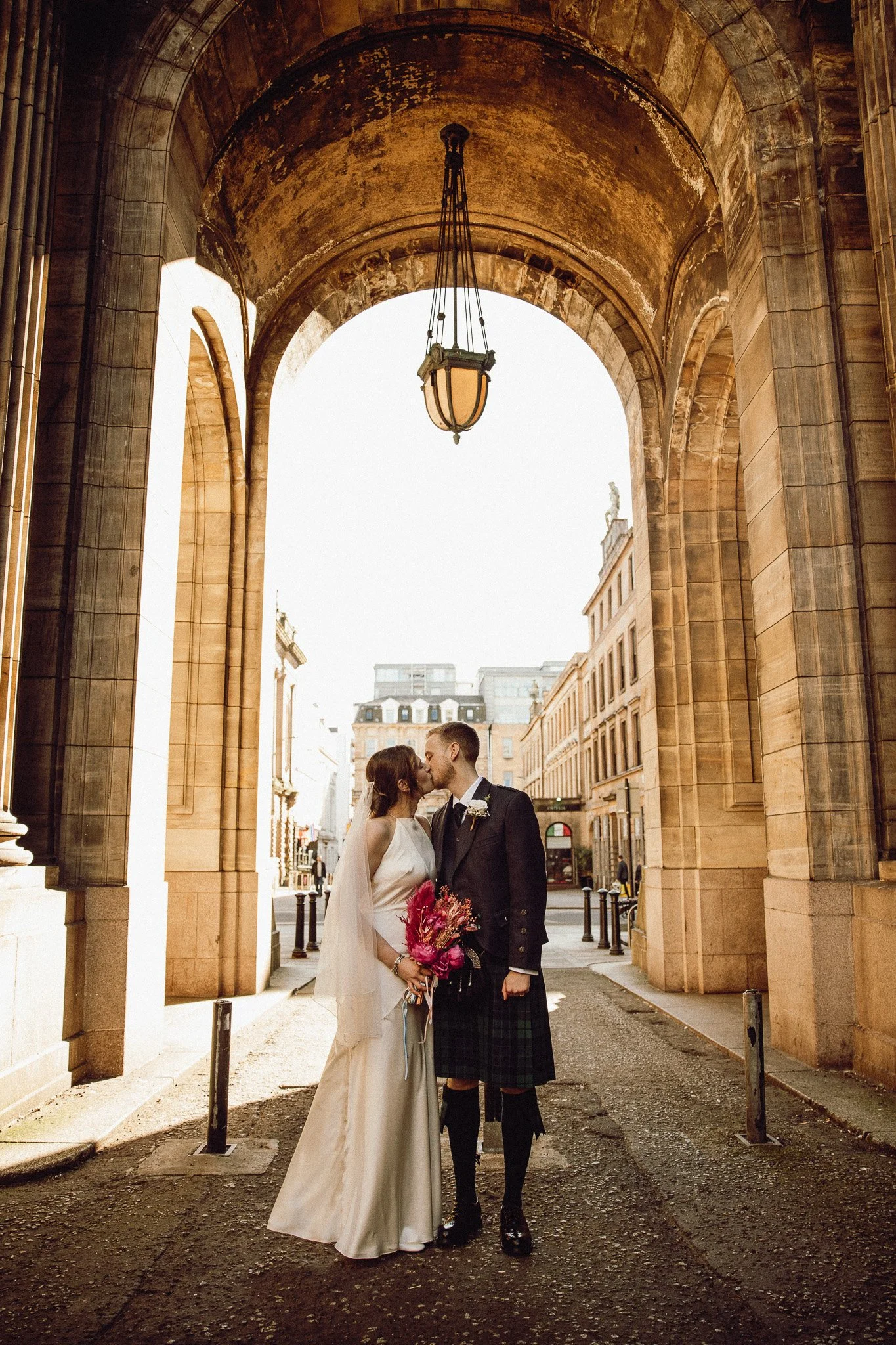 A bride and groom sharing a kiss under a large archway in an urban setting, with old stone buildings and a street in the background, the bride holding a bouquet of pink flowers.