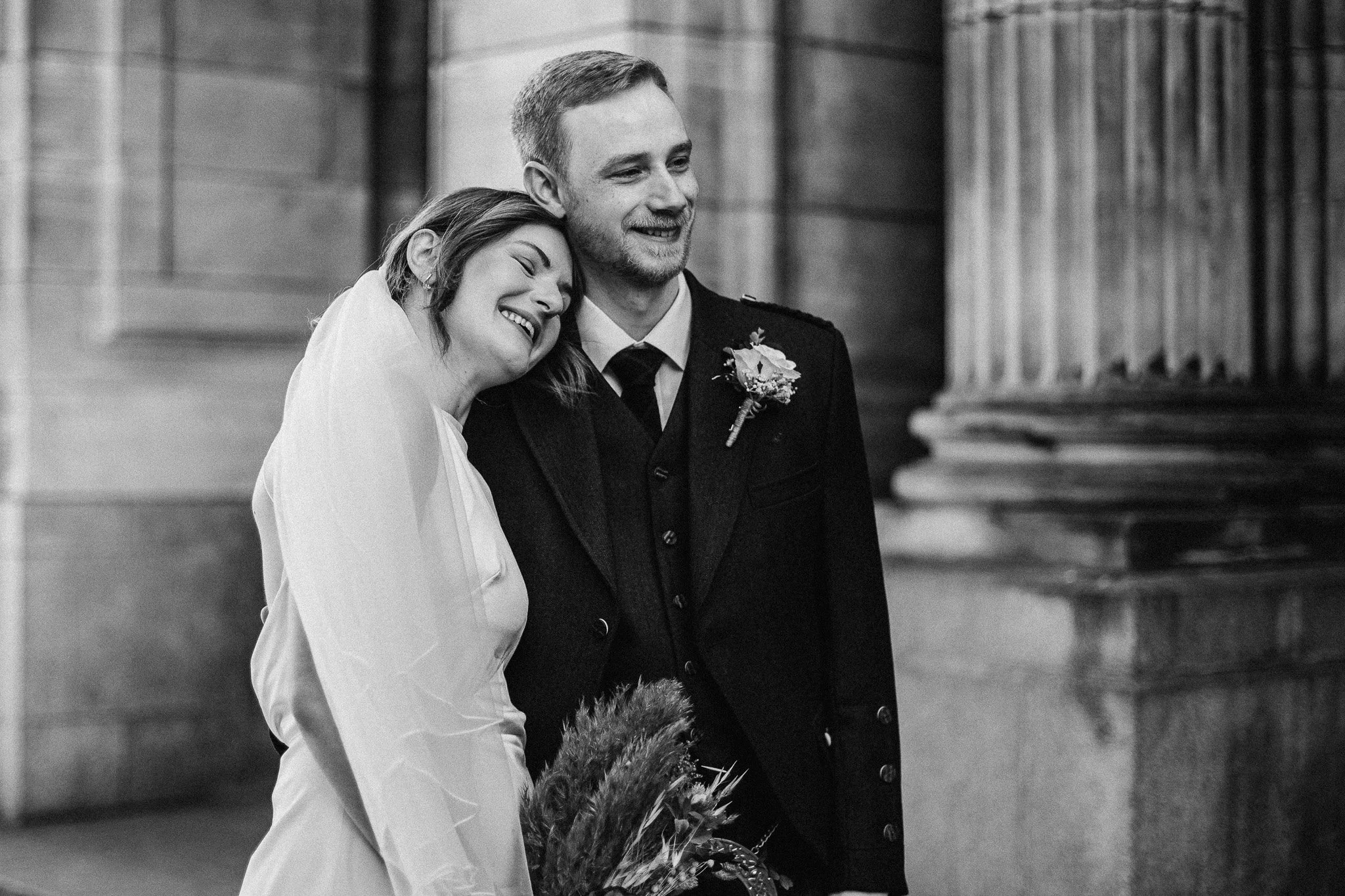 A black and white photo of a smiling bride and groom, with the bride resting her head on the groom's shoulder, standing near large stone columns.