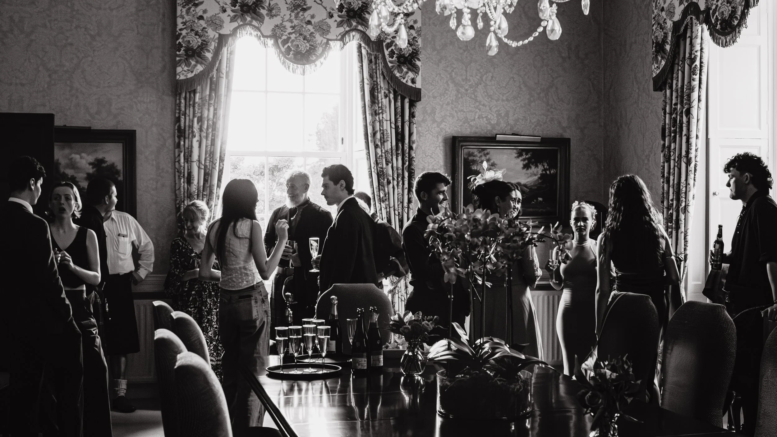 People attending a social gathering or party inside a well-decorated room with curtains and chandelier, holding drinks and engaging in conversations.
