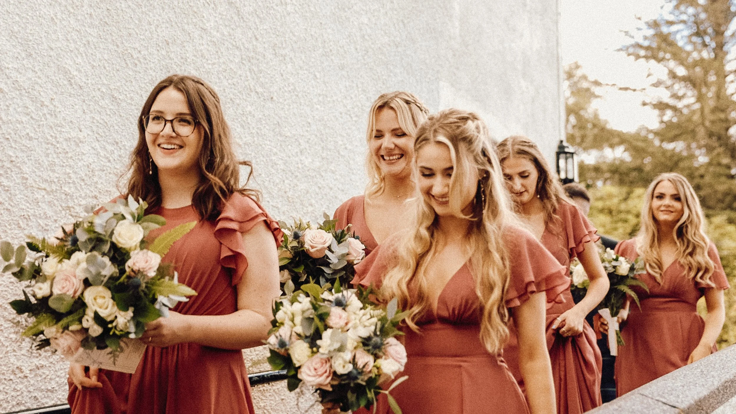 A group of women in matching rust-colored dresses holding bouquets of flowers walking outdoors.