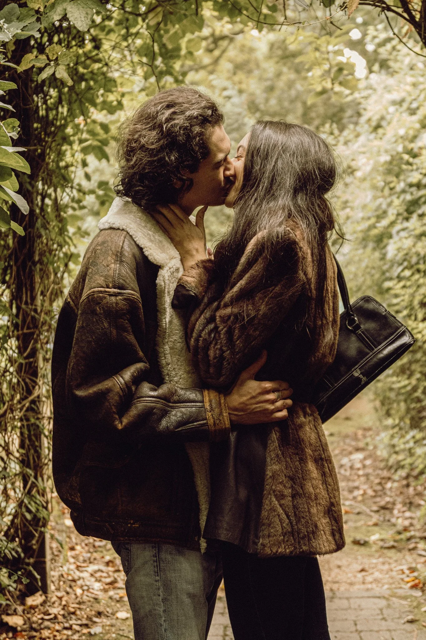 A couple kissing on a forest path surrounded by greenery and fallen leaves, dressed in warm jackets.