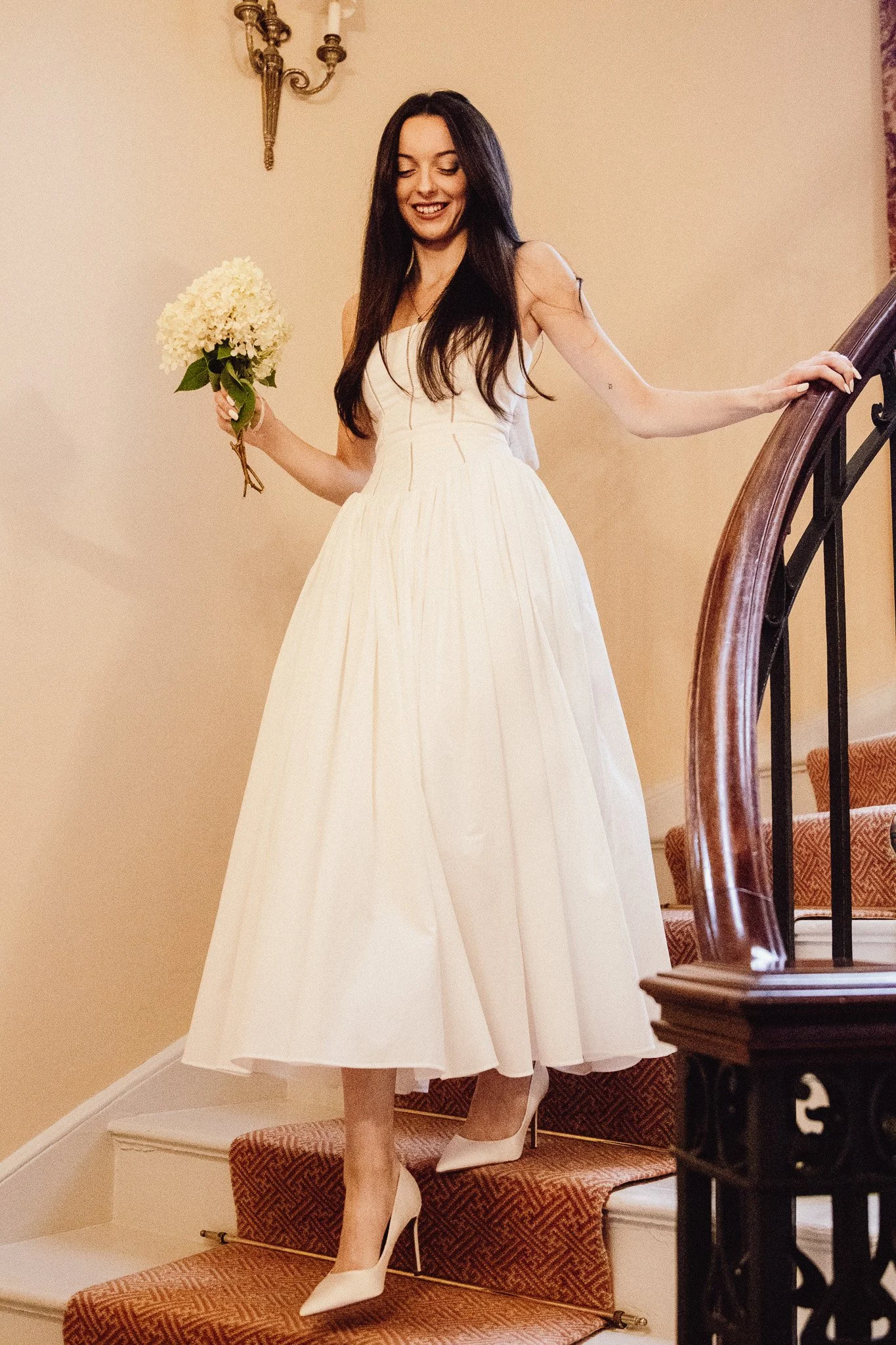 A woman in a white dress and high heels is descending a staircase, holding a bouquet of white flowers, smiling, with long dark hair, in an indoor setting.