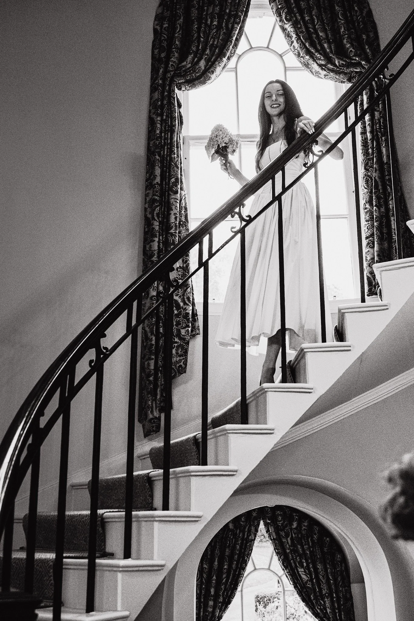 A woman in a white dress standing on a staircase, holding a bouquet of flowers, with a large window and ornate curtains in the background.