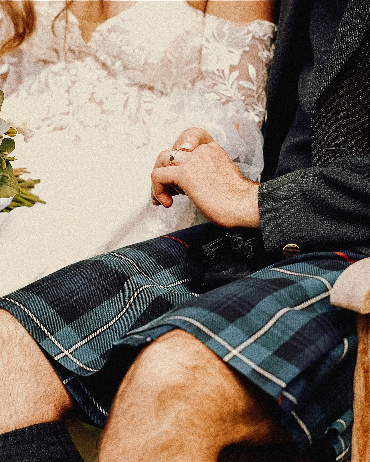 Close-up of a couple holding hands during a wedding, with the bride in a white lace dress and the groom in a dark blazer and plaid kilt.