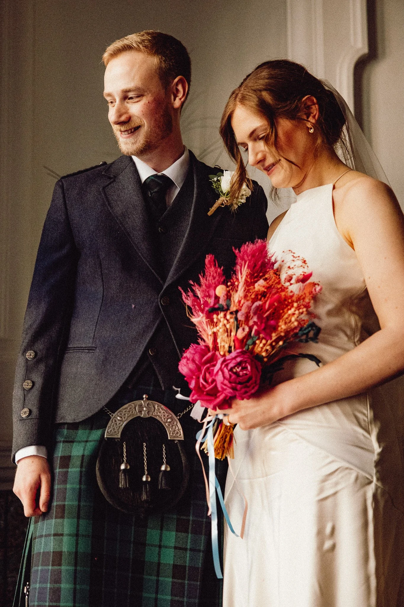 A bride and groom at their wedding, with the bride holding a bouquet of vibrant pink, orange, and red flowers, and the groom wearing a traditional Scottish kilt and jacket.
