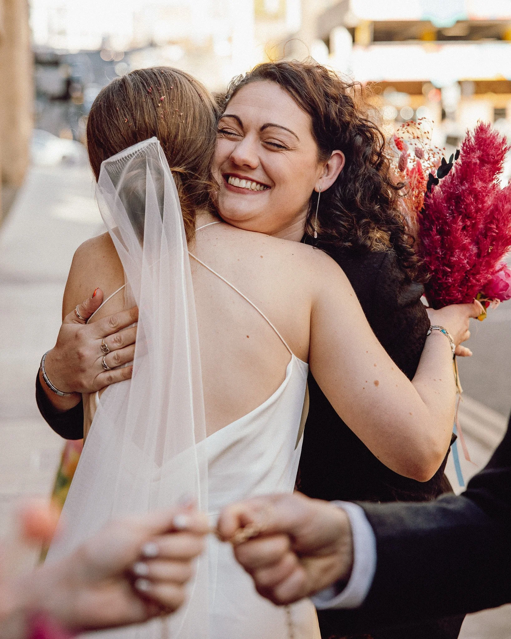 Two women hugging, one in a wedding dress and veil, the other holding a bouquet, during a wedding celebration.