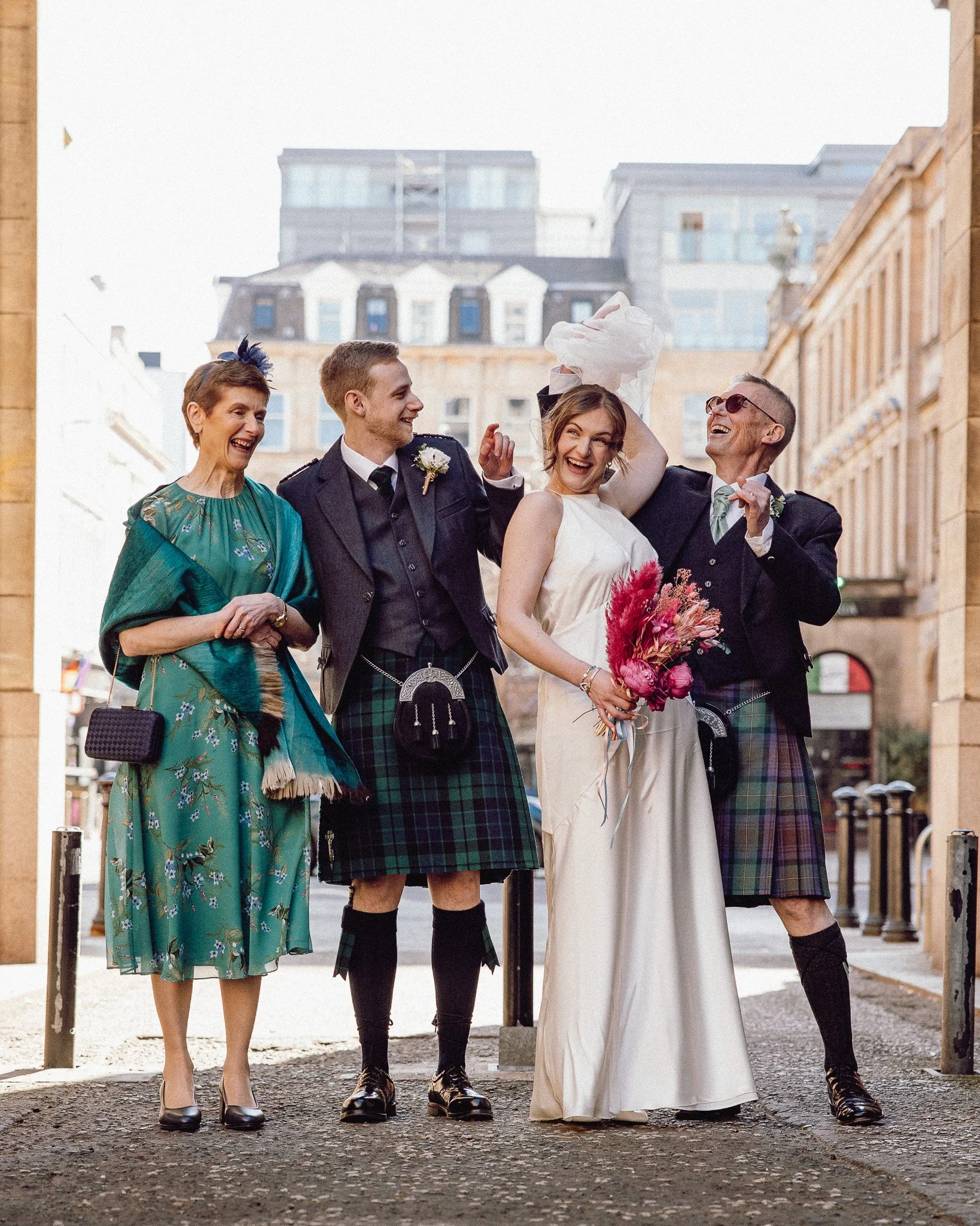 A group of five people, dressed in wedding attire including kilts, standing outdoors on a city street, all laughing and celebrating. The woman in the center is holding a bouquet of flowers.