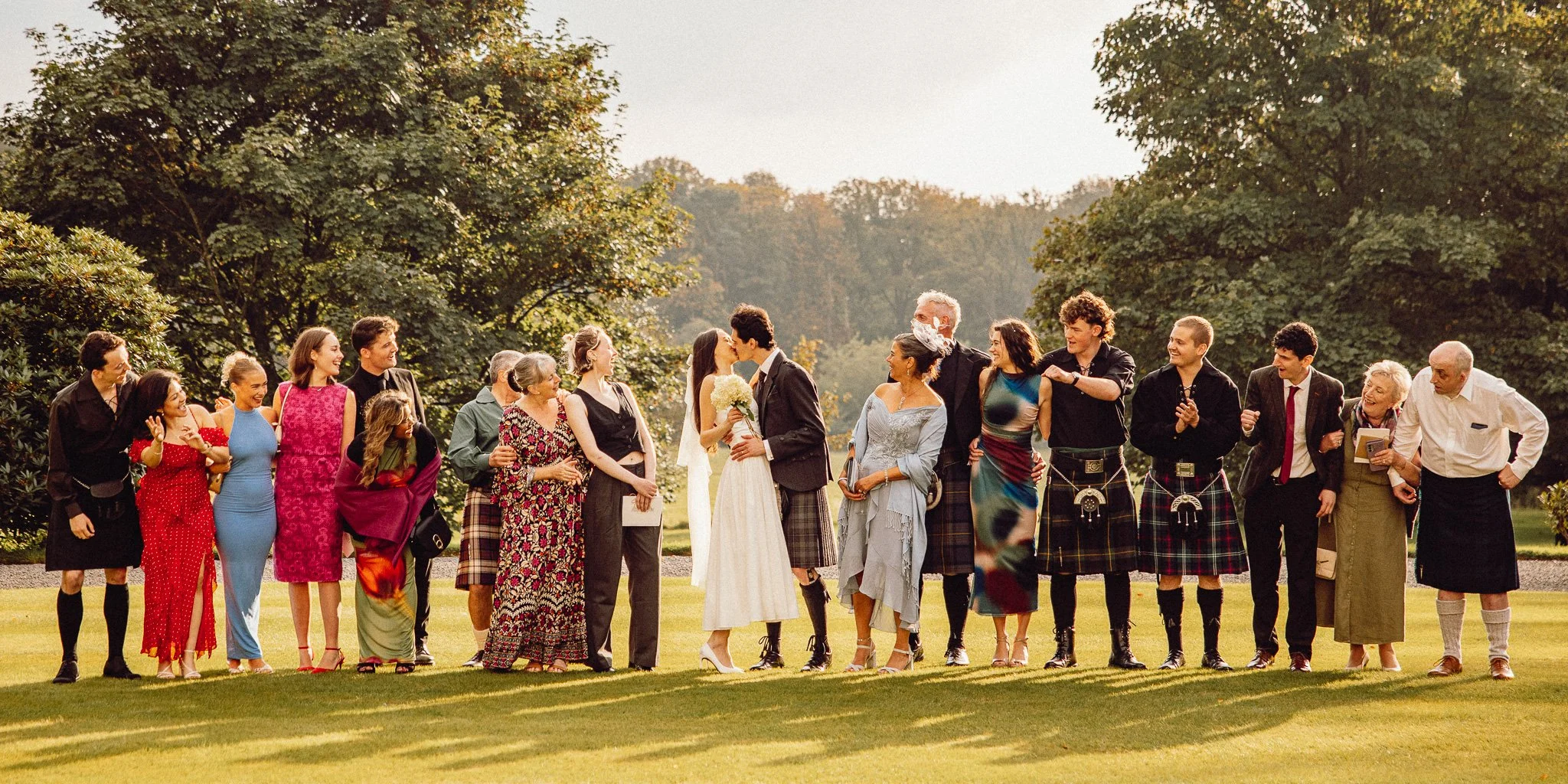 A group of people at a wedding, with the bride and groom kissing in the center, surrounded by family and friends on a lawn with trees in the background.