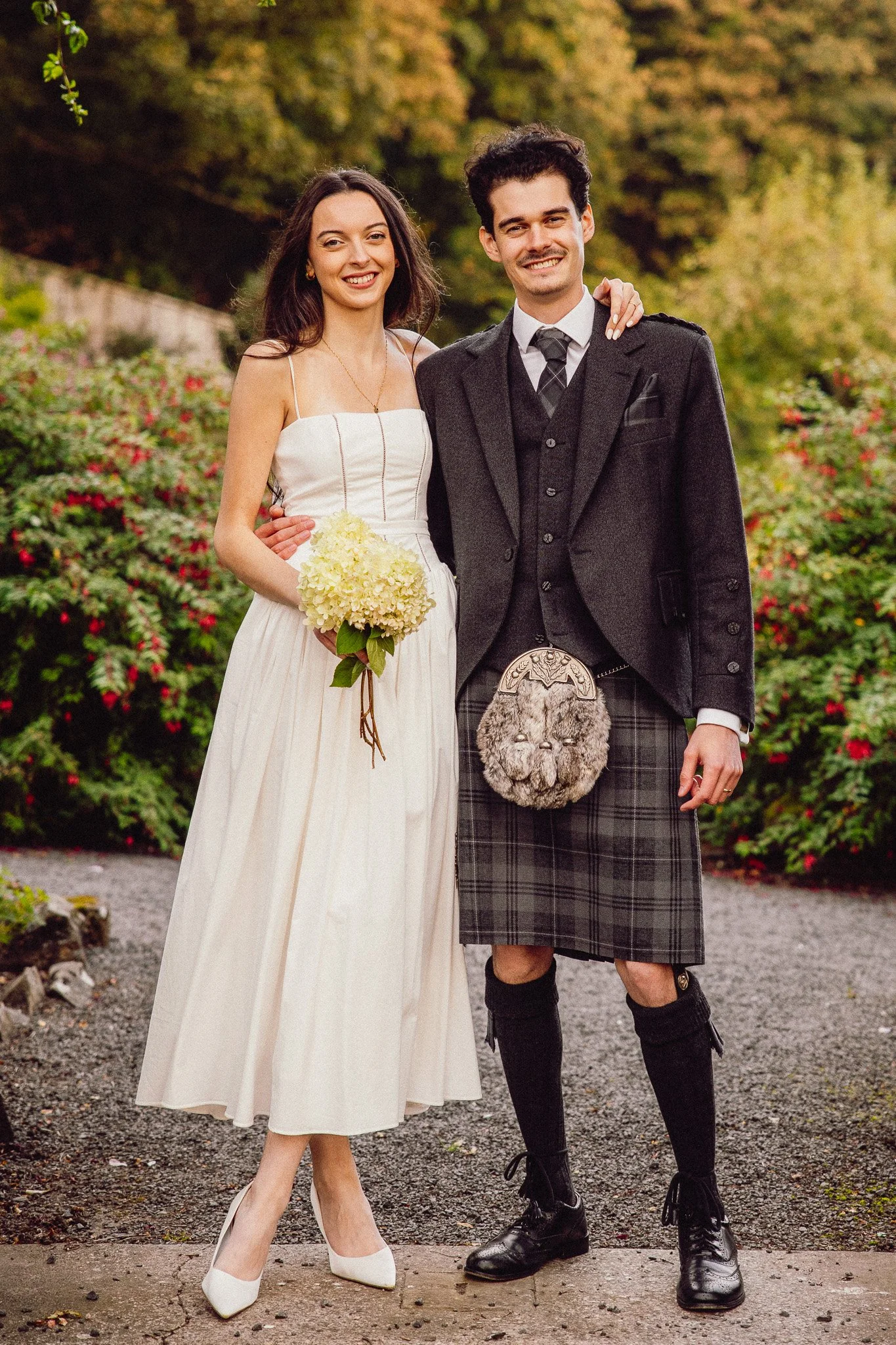 A newlywed couple standing outdoors, smiling at the camera. The woman is wearing a white wedding dress and holding a bouquet of white flowers, and the man is dressed in traditional Scottish attire, including a kilt and a sporran. The background is fi