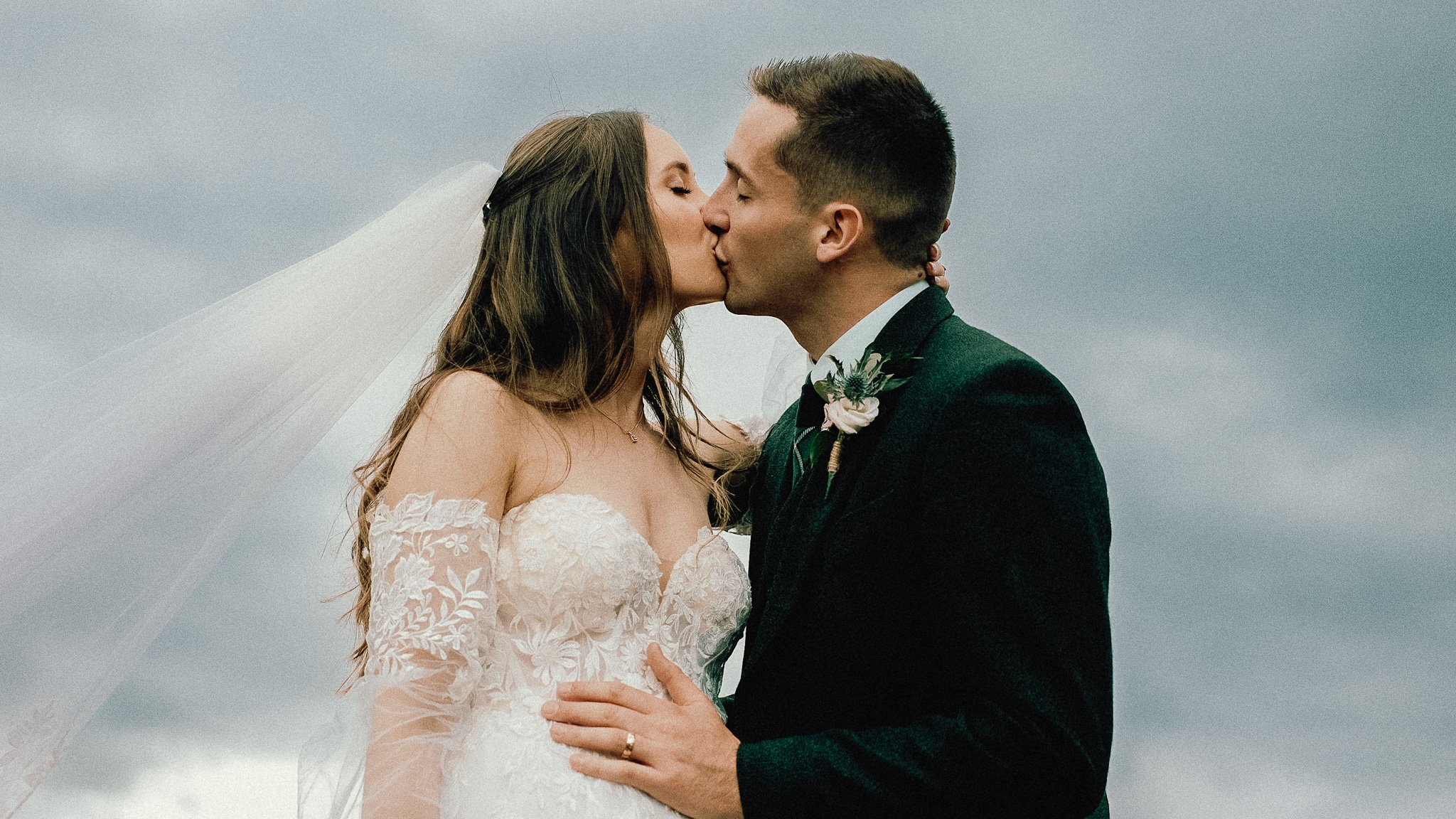 Bride and groom kissing outdoors on cloudy day, bride in lace wedding dress with veil, groom in dark suit with boutonniere, bride's hand on groom's chest, groom wearing wedding ring.