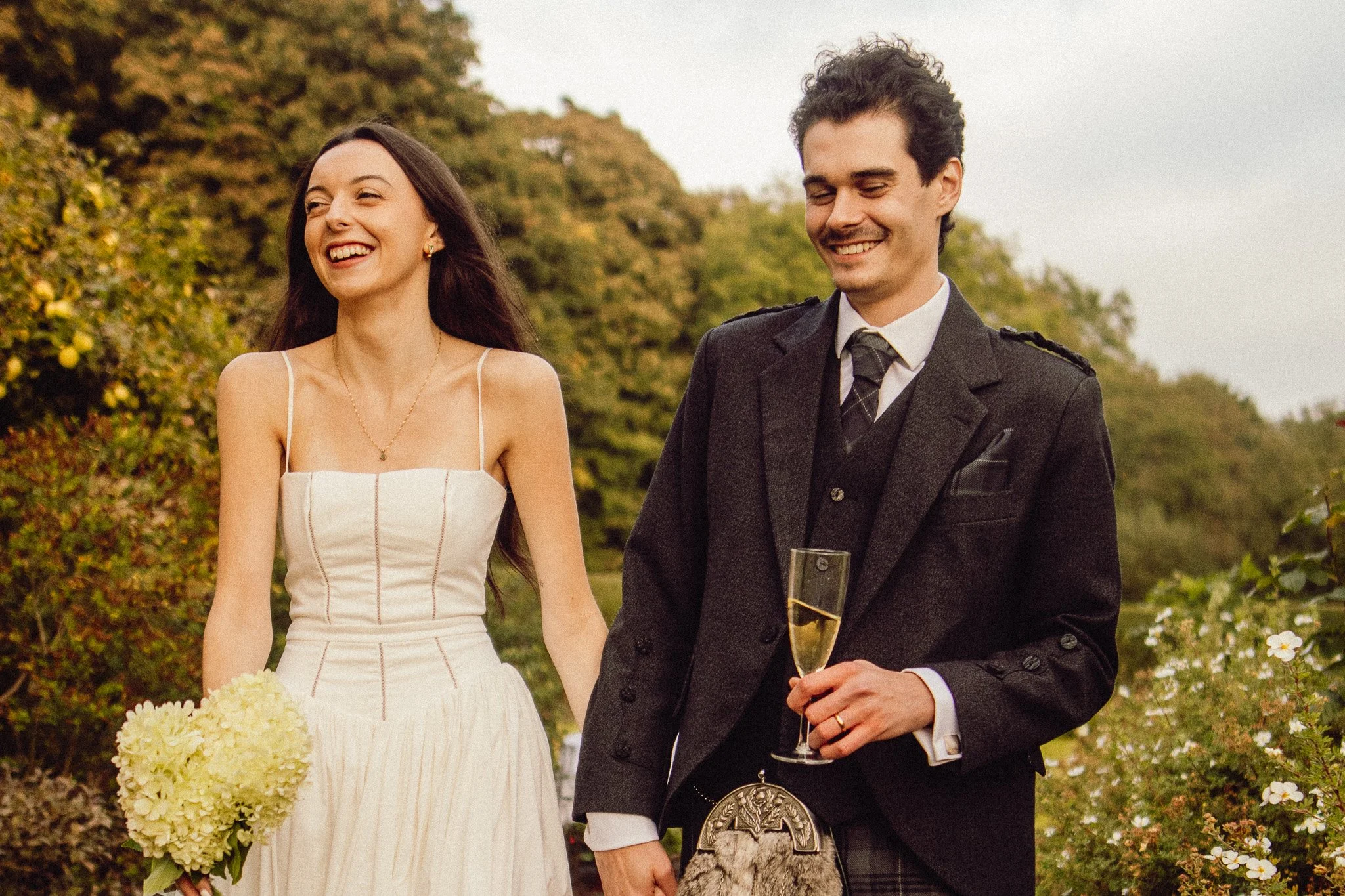 A smiling woman in a white dress holding a bouquet, walking outdoors with a smiling man in a dark suit holding a champagne glass, in a lush garden setting.