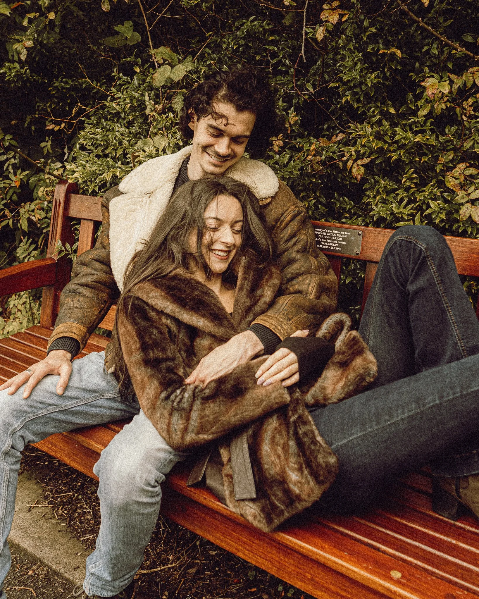 A young couple sitting on a park bench, smiling and looking at a smartphone together. The man is behind the woman, with his arms around her. They are surrounded by green foliage.