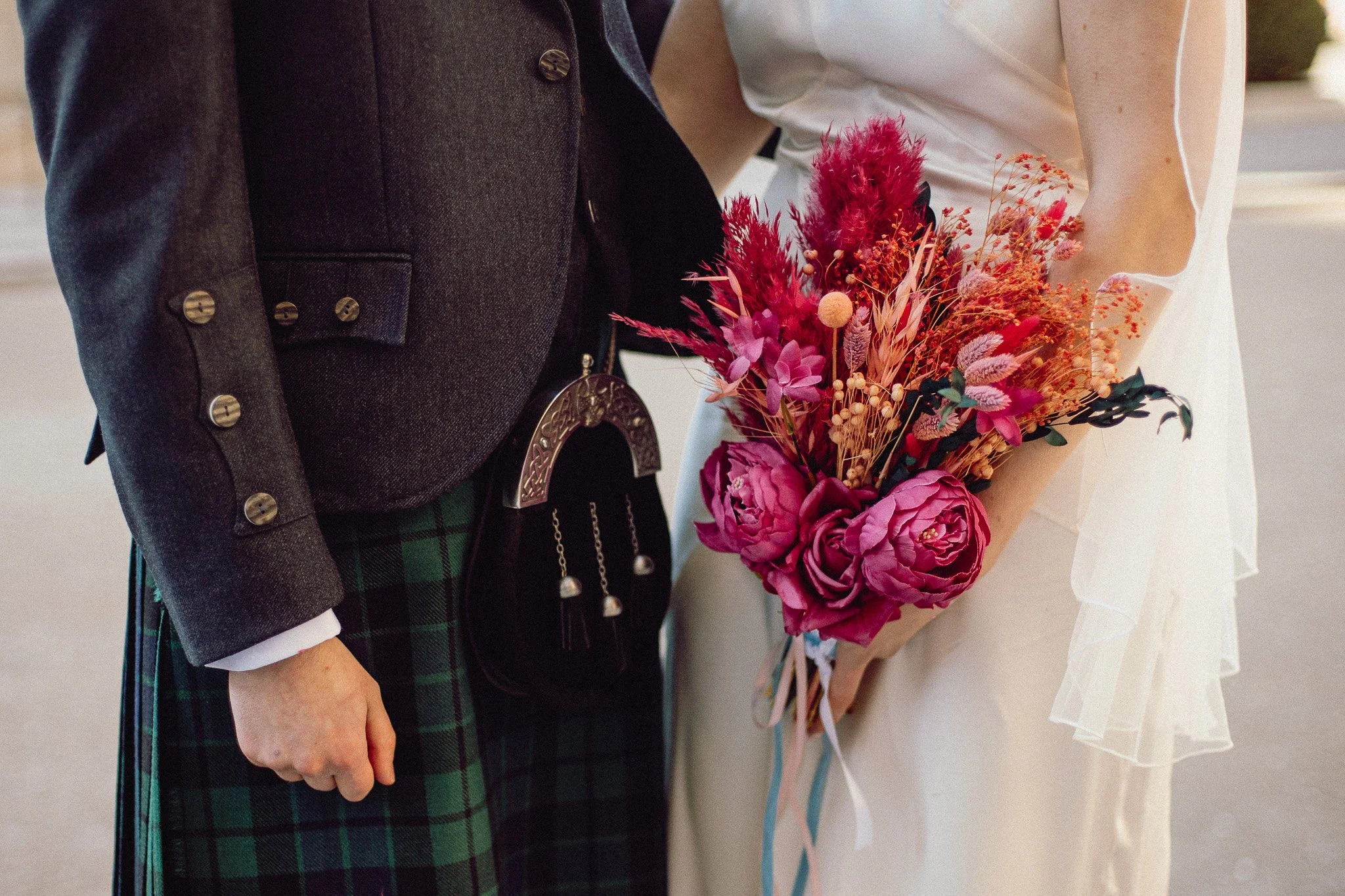 Close-up of a bride holding a bouquet of pink and red flowers next to a groom in formal attire, with part of his kilt visible.