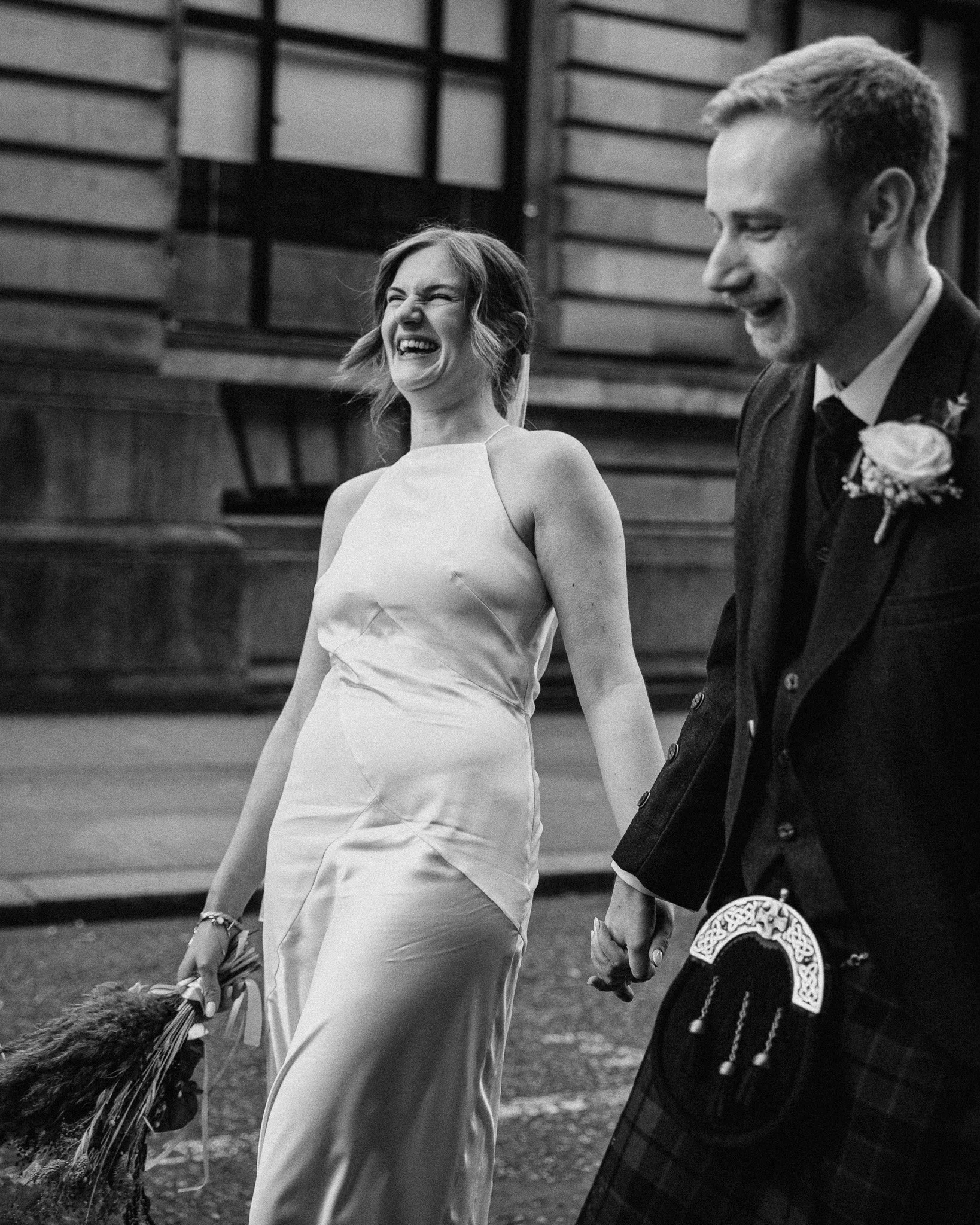 A smiling woman in a wedding dress holding a bouquet walking hand in hand with a man dressed in a suit and kilt, both enjoying a moment together.