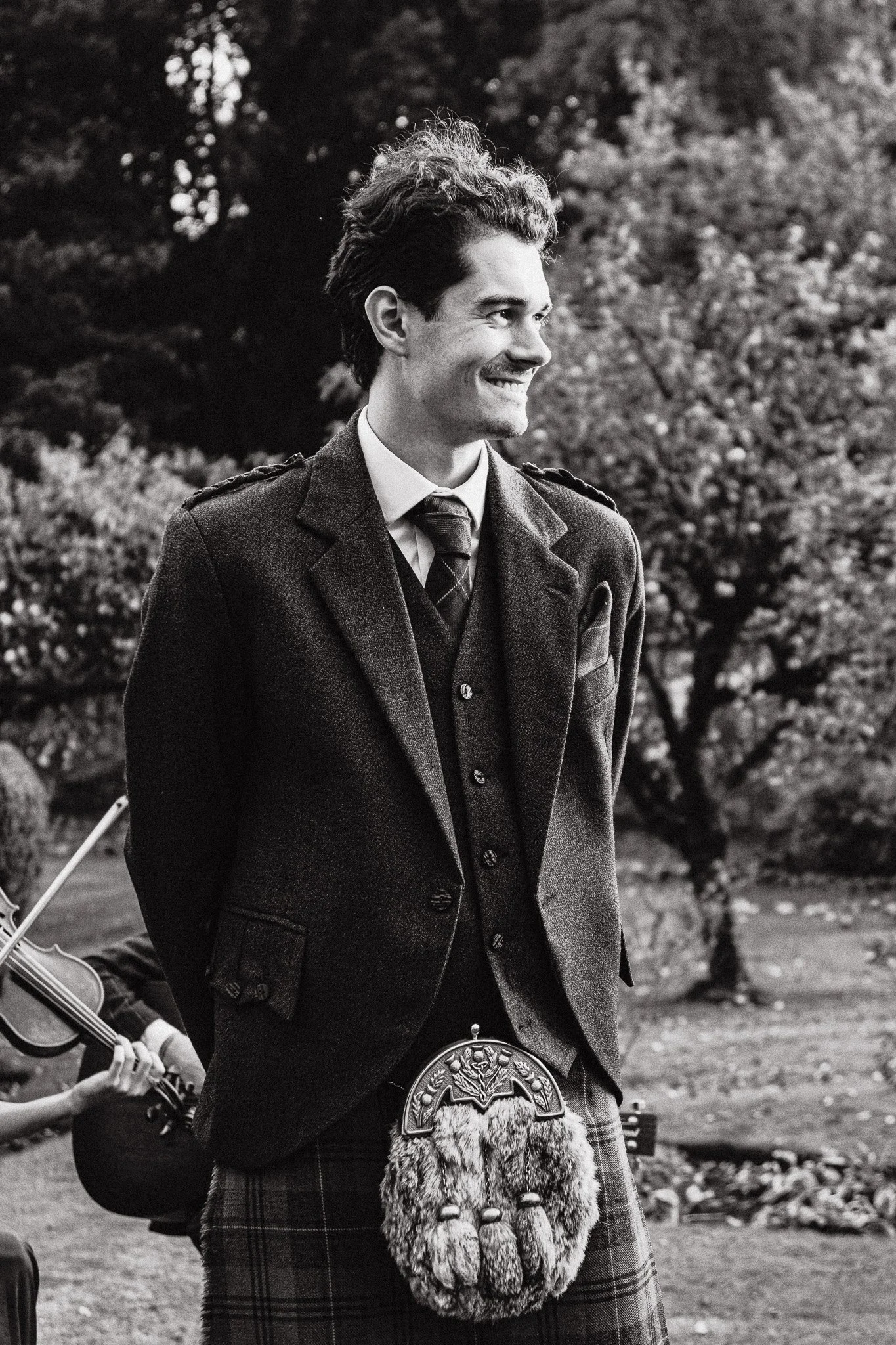 A young man in traditional Scottish attire, including a kilt and a sporran, standing outdoors with trees in the background, smiling.