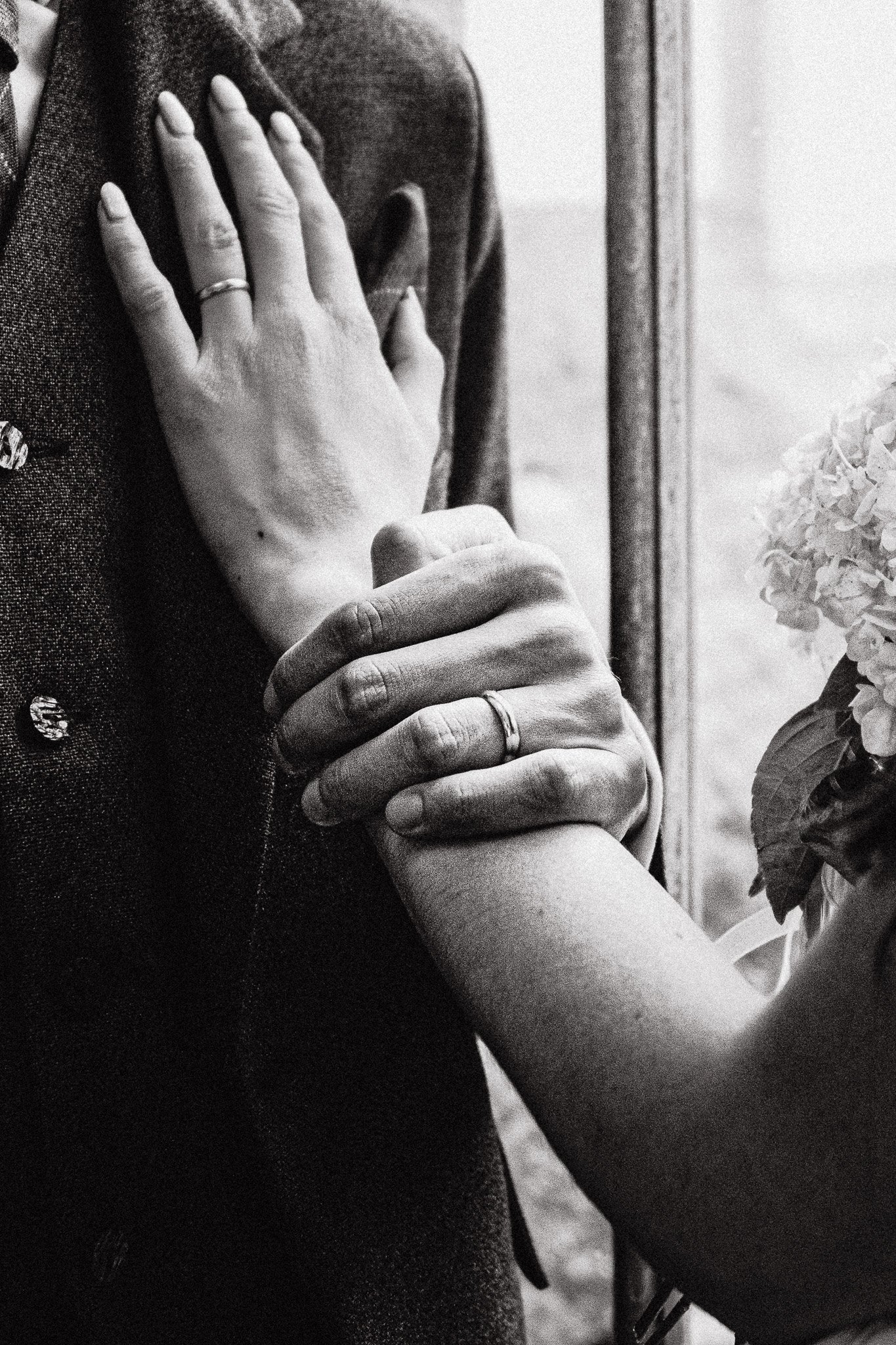 Close-up of a couple holding hands, with wedding rings, next to a window with a flower bouquet nearby, in black and white.