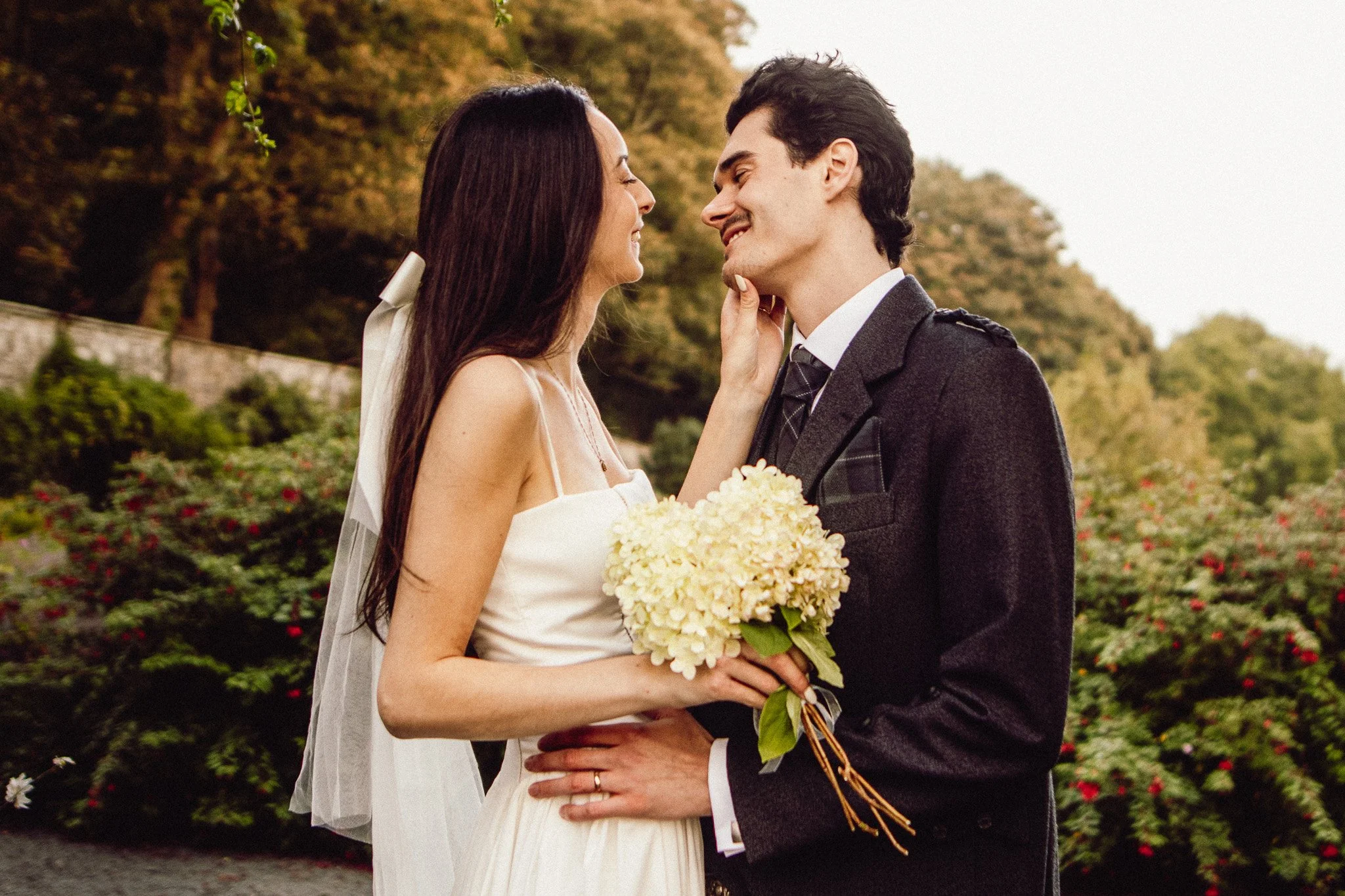 A newlywed couple standing close together outdoors, smiling and looking into each other's eyes. The bride is holding a bouquet of white flowers and is touching the groom's face. They are surrounded by greenery and flowers in the background.