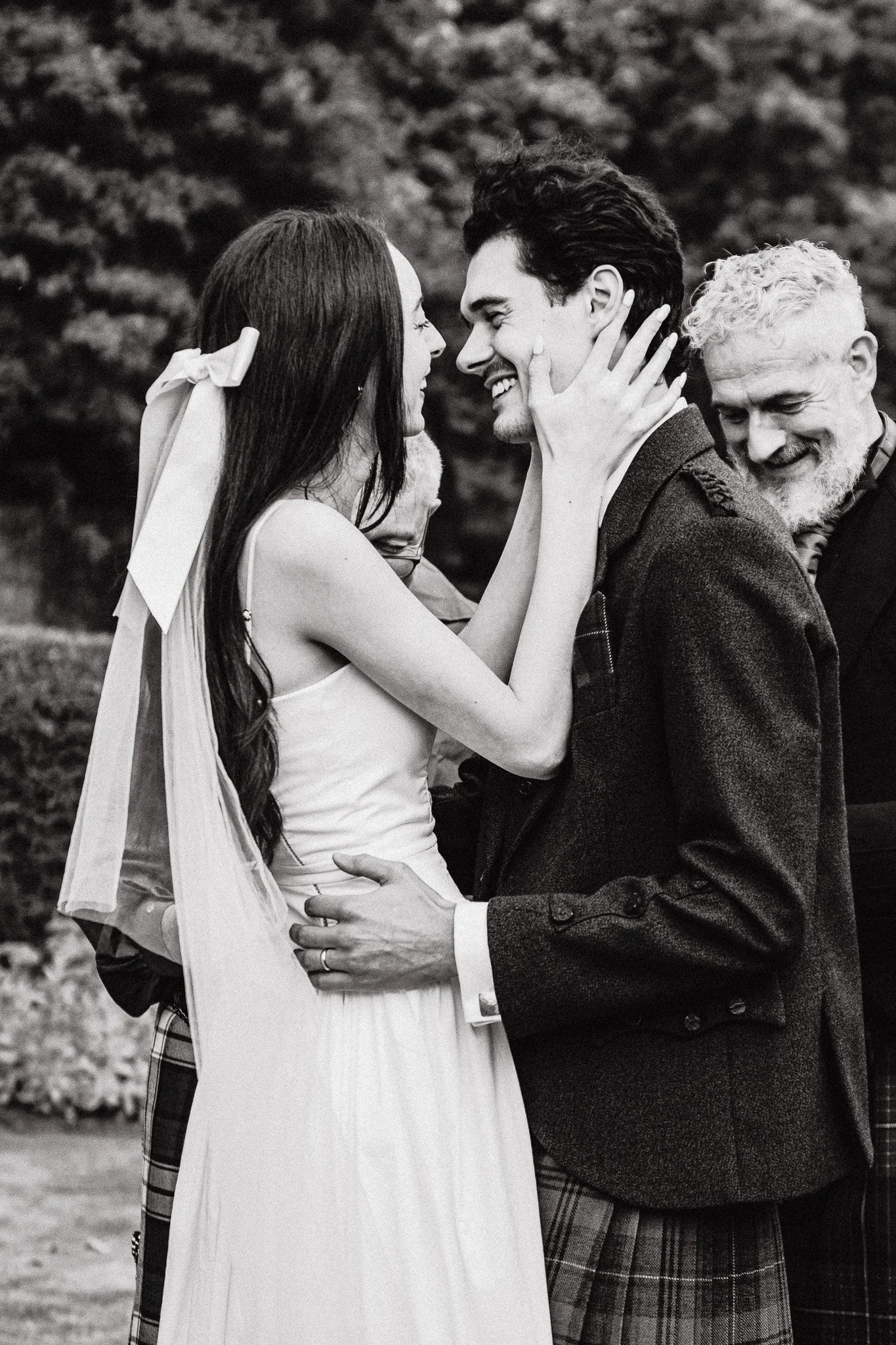 A black and white photo of a couple kissing, with an older man smiling behind them, outdoors with trees in the background.