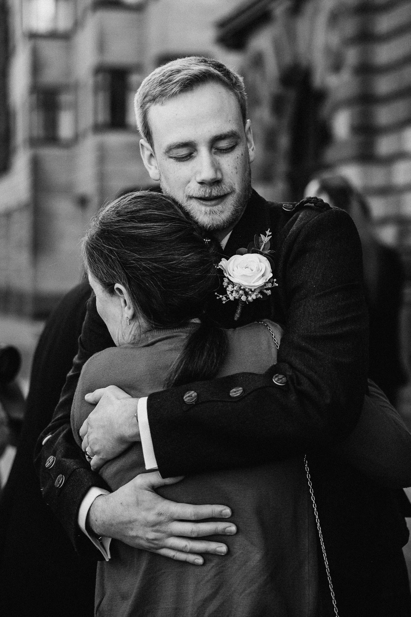 A young man, wearing a suit with a boutonnière, embraces a woman with long hair in a hug in an outdoor urban setting.