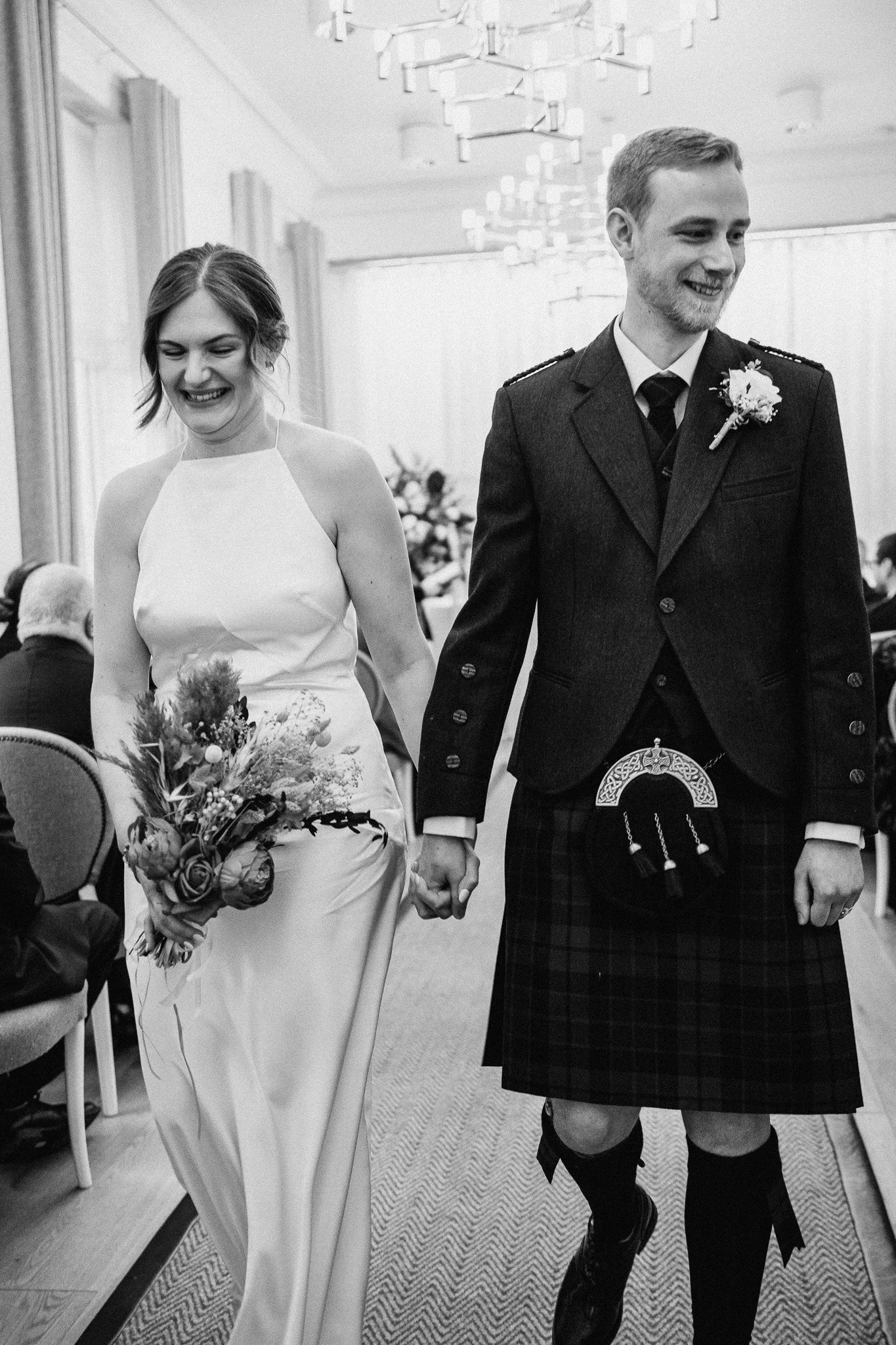 A bride and groom walking down the aisle at their wedding ceremony, holding hands and smiling. The bride is wearing a sleeveless wedding gown and carrying a bouquet, while the groom is in a traditional Scottish kilt and jacket.