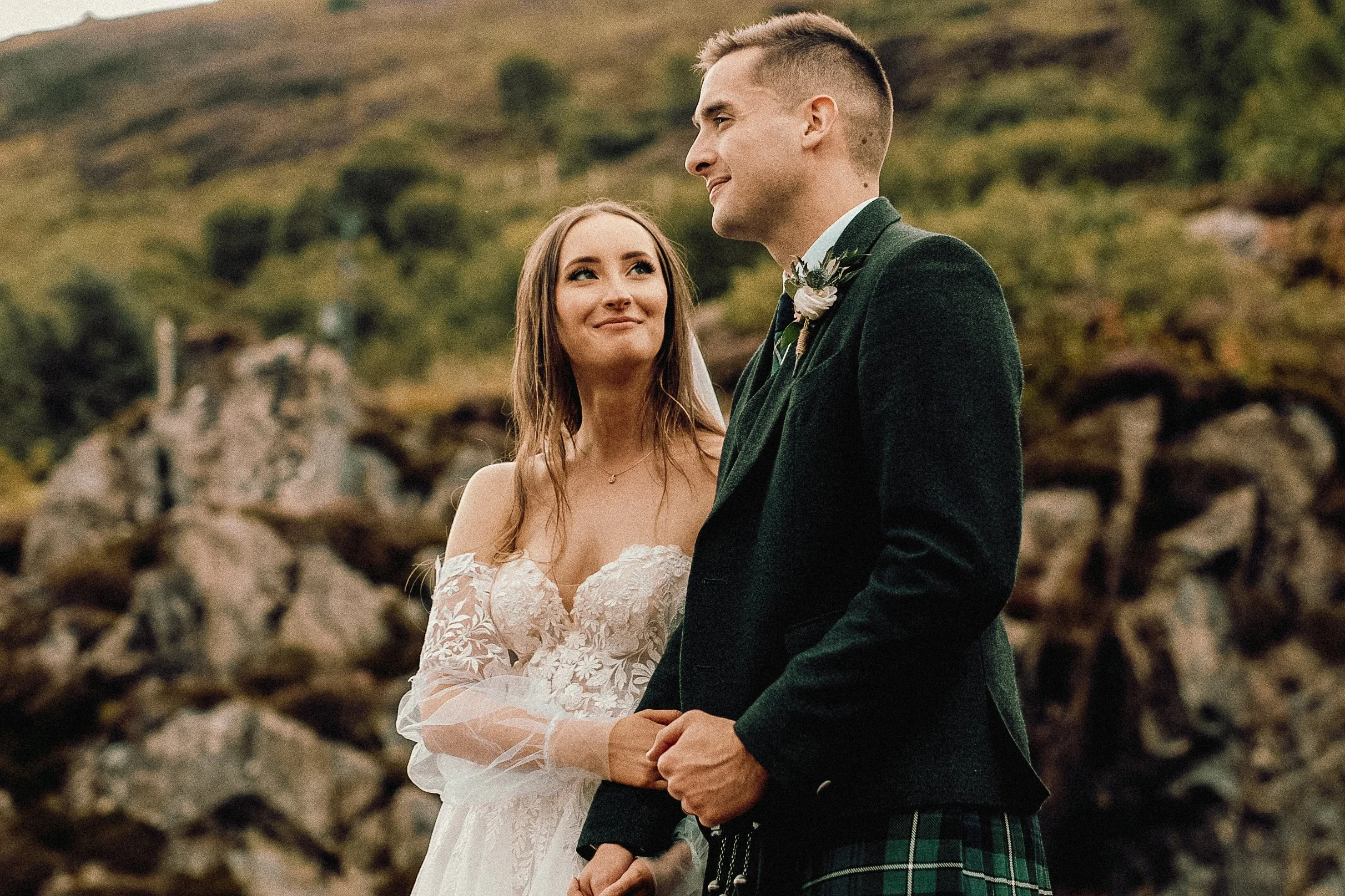 A bride and groom holding hands during their outdoor wedding ceremony, with green hills and rocks in the background, and both dressed in wedding attire.