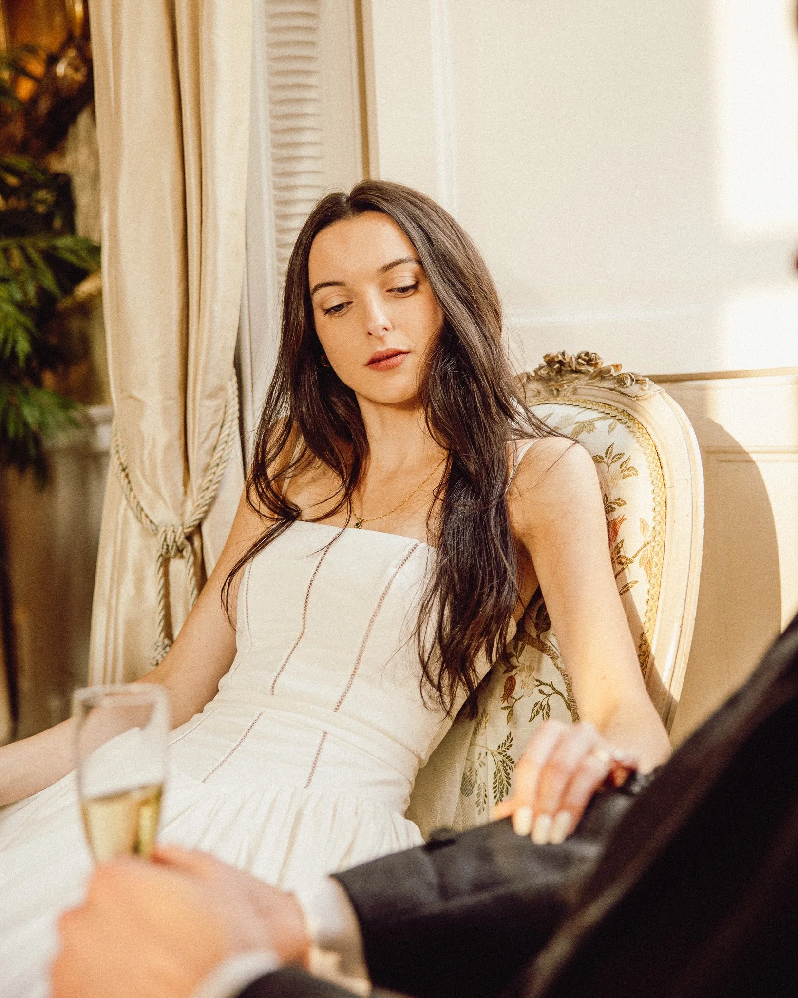 Young woman in a white dress sitting on a vintage chair, with a drink in front of her, in a well-lit room with golden curtains and a plant in the background.