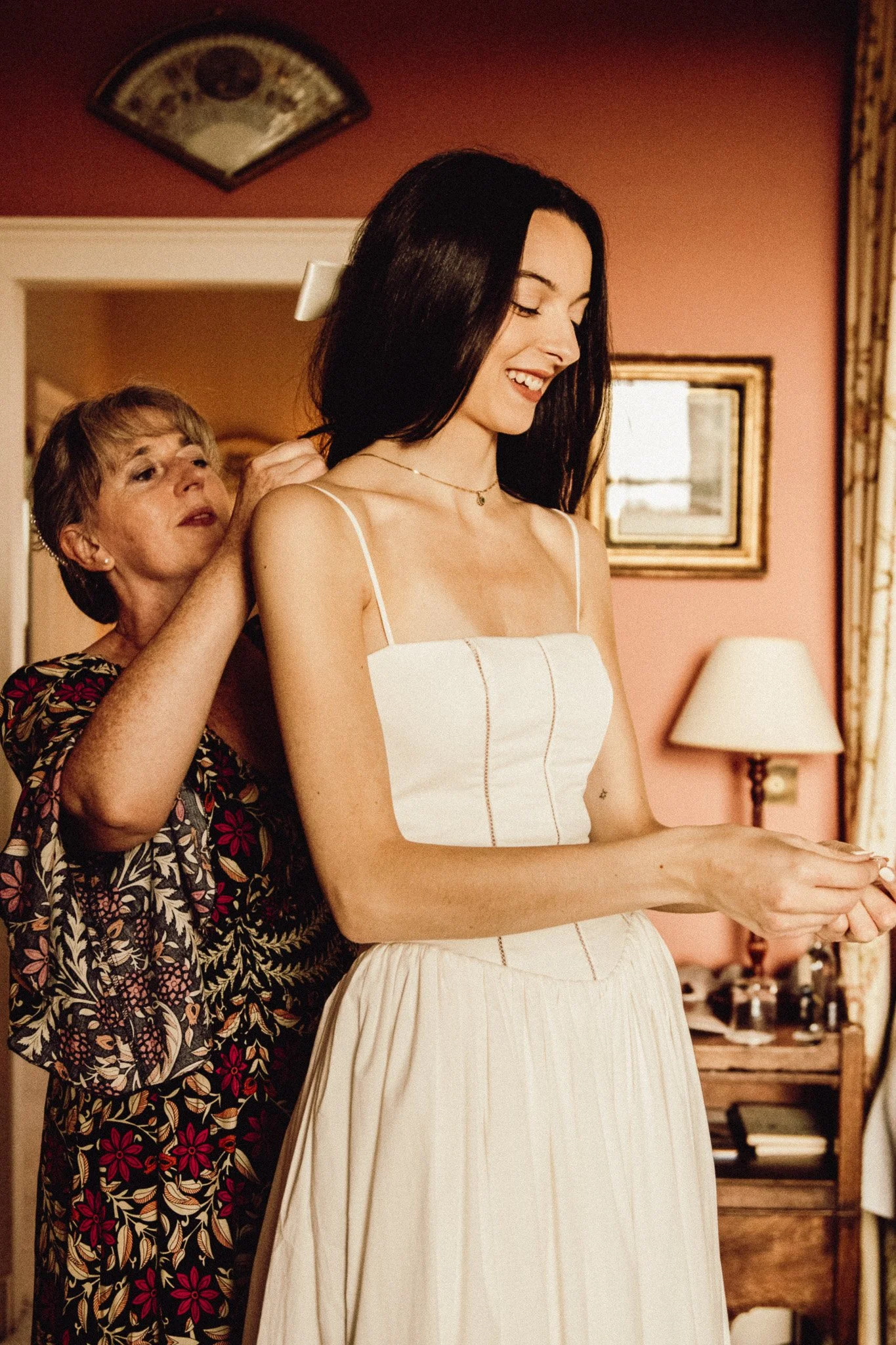 A young woman in a white dress has her hair prepared by an older woman in a floral blouse in a warmly decorated room.