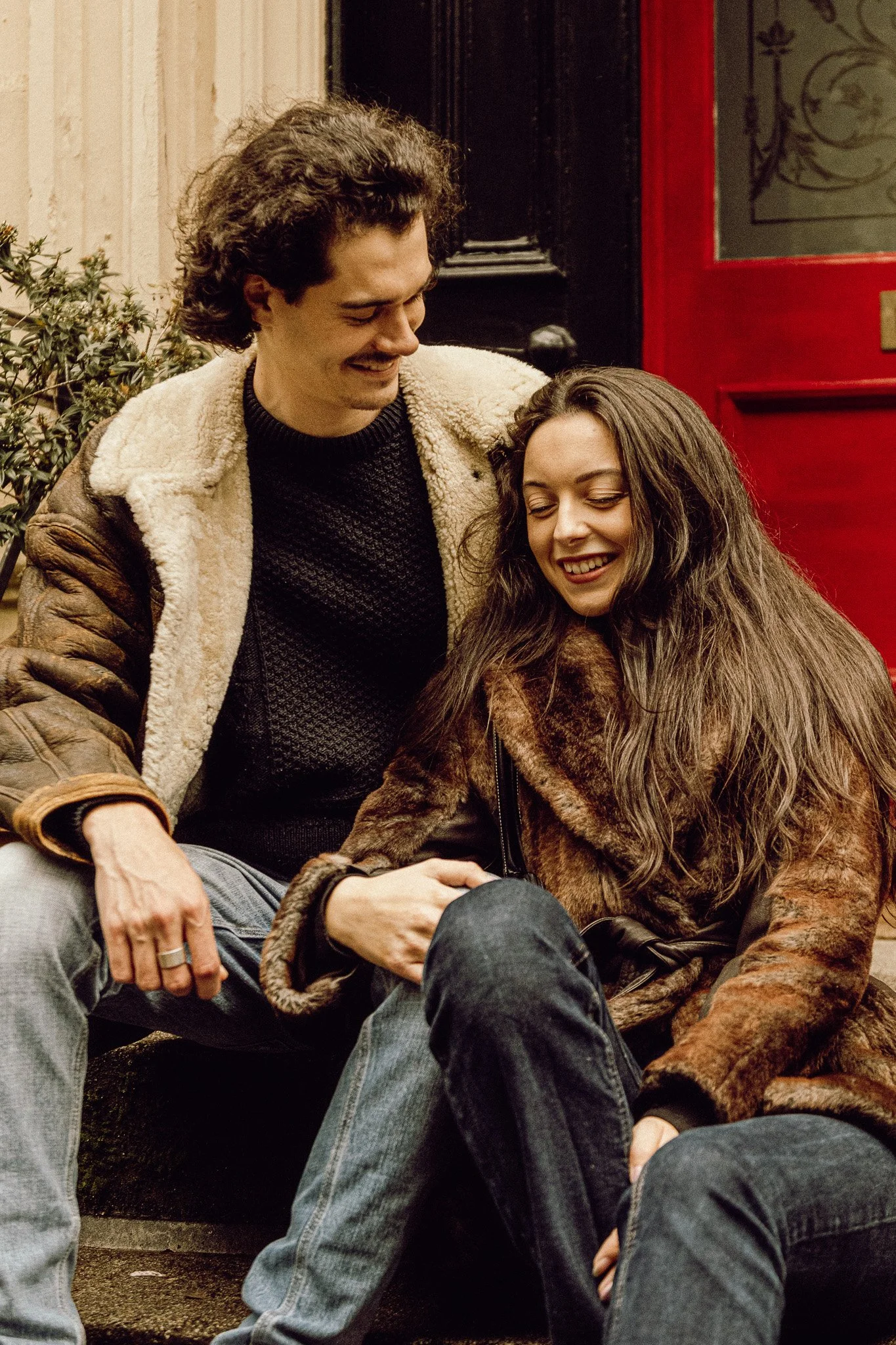 A young man and woman sitting on the steps outside a building, smiling and looking at each other. The man has curly dark hair and wears a brown jacket with a shearling collar. The woman has long, wavy brown hair and wears a fur coat. They appear to b